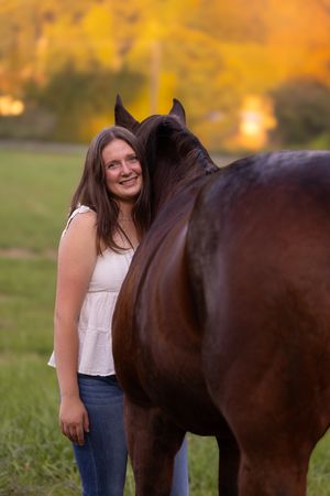 A person in a white top and jeans stands beside a dark brown horse in a sunlit field at golden hour.