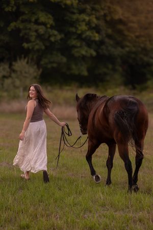 A graceful figure in a flowing dress walks through a grassy field alongside a dark brown horse at sunset.