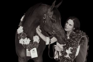 Black and white photo of a smiling woman with a decorated horse wearing floral garlands against a dark background.