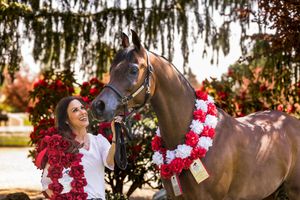Woman smiling beside a decorated horse wearing red and white floral garlands outdoors.
