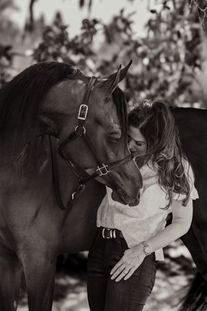 Black and white photo of a woman tenderly nuzzling a dark horse wearing a bridle outdoors.
