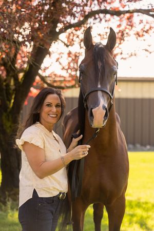 Smiling woman in yellow blouse poses with a bay horse outdoors near a flowering tree in warm sunlight.