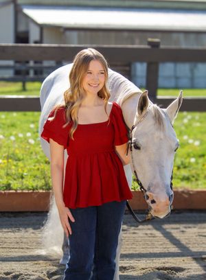 A woman in a red top and navy pants poses with a white horse in a sunny outdoor paddock area.