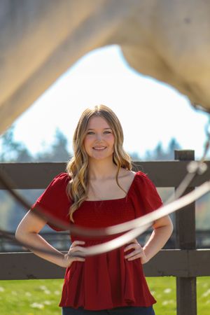 A person in a red off-shoulder dress poses outdoors against a wooden railing on a sunny day.