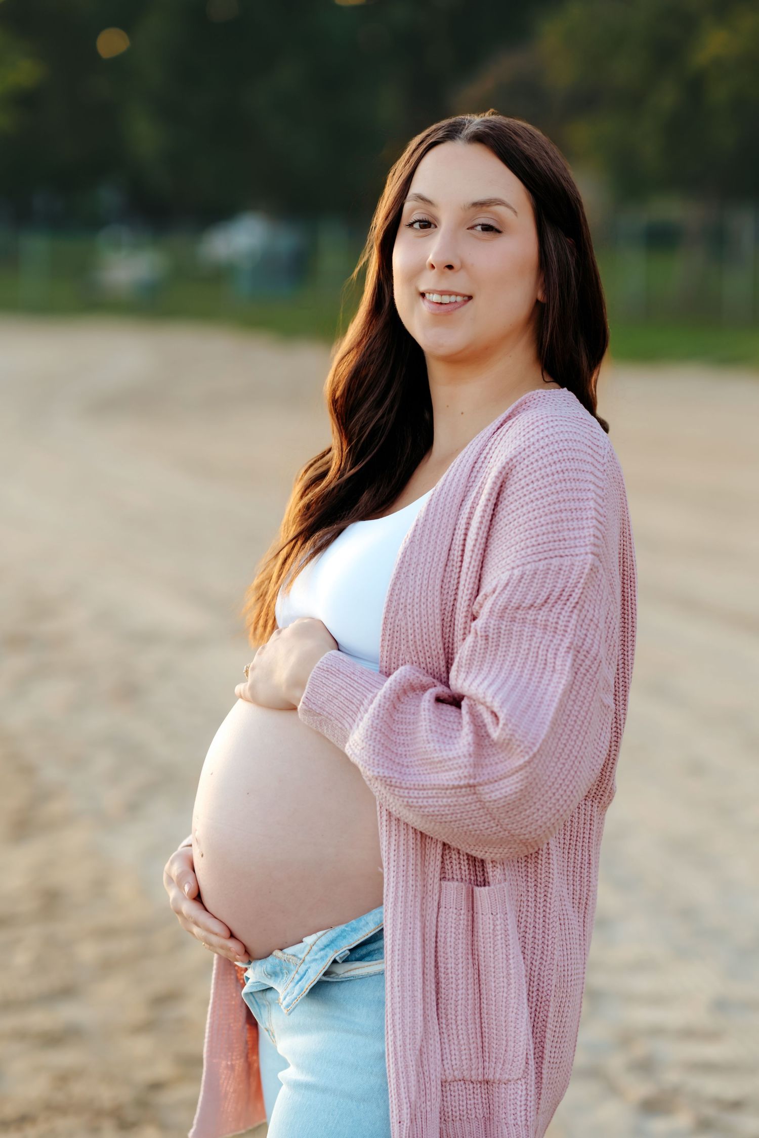 A maternity photo session on the beach shows someone in a white top and pink cardigan posing at sunset.