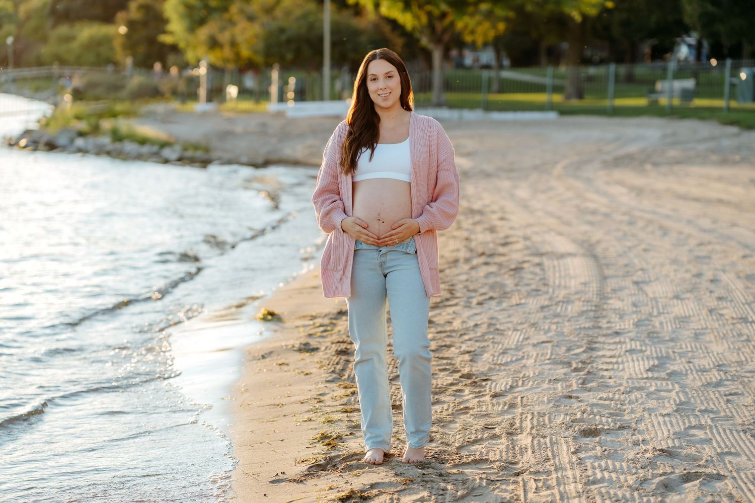 Woman walking barefoot along a sandy beach shoreline at sunset wearing light blue jeans and pink top.