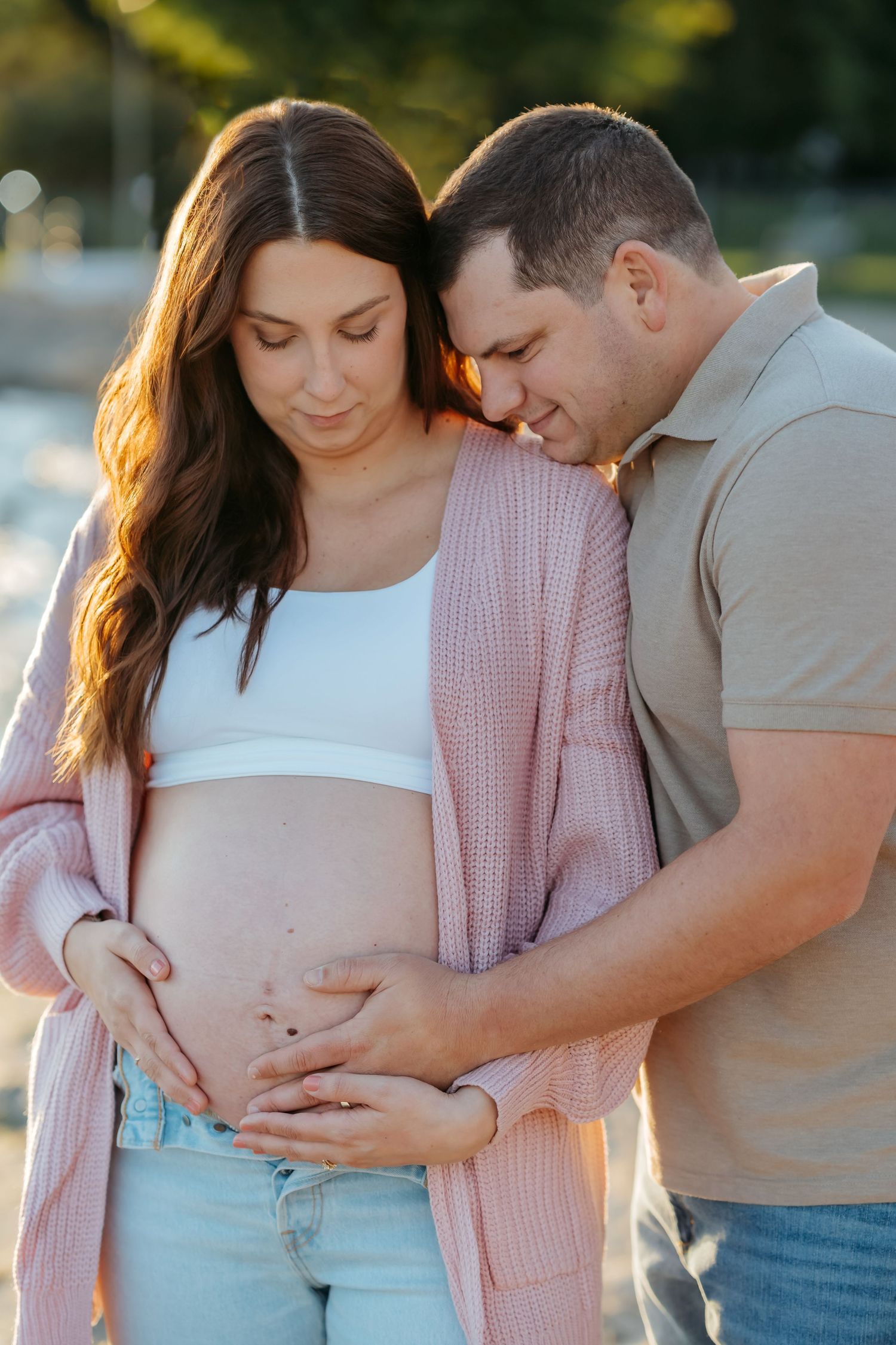 A couple shares an intimate moment during a maternity photoshoot at sunset in a park setting.