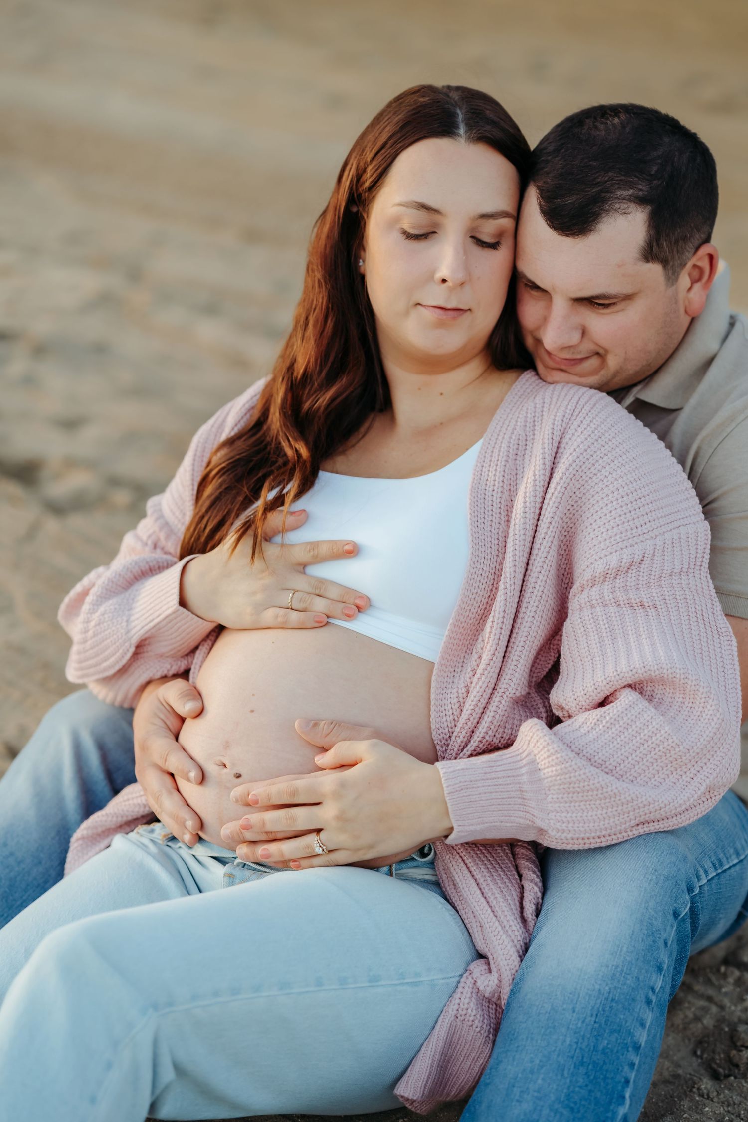 Couple in casual attire sharing tender moments during maternity photoshoot on sandy beach at sunset.
