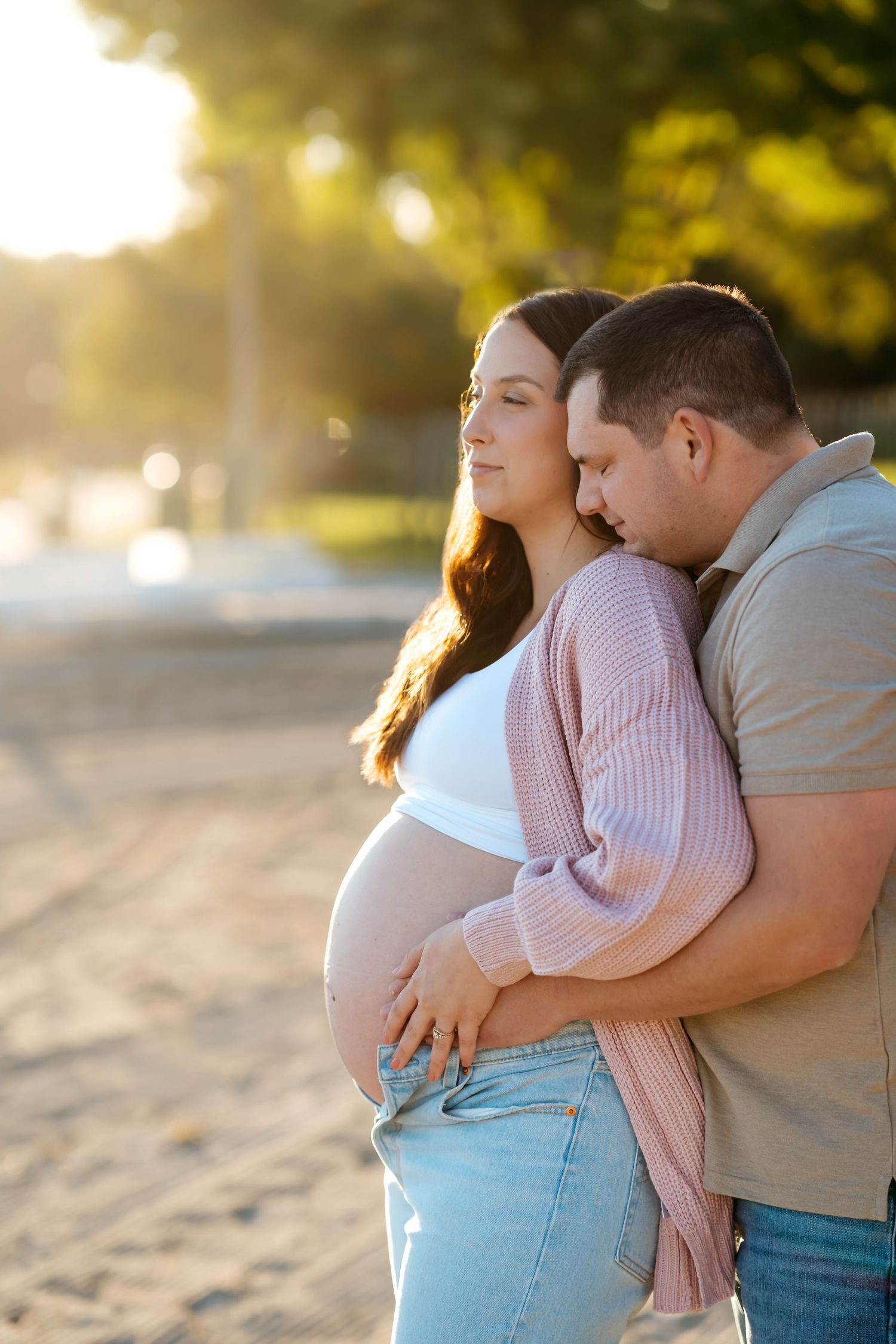Couple embraces on sandy beach during golden hour sunset in romantic maternity photoshoot.