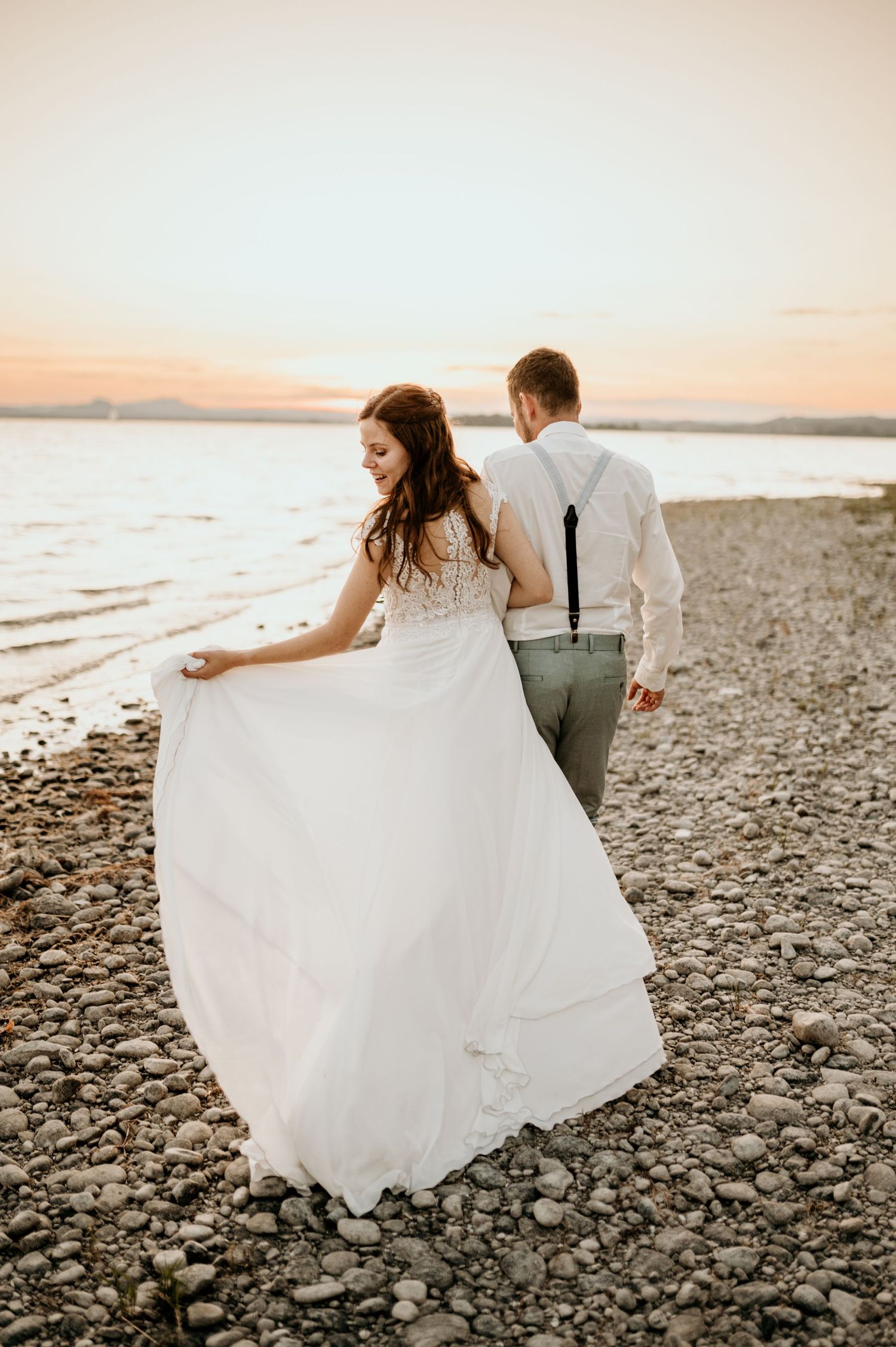 Couple walking along rocky beach at sunset with flowing wedding dress.