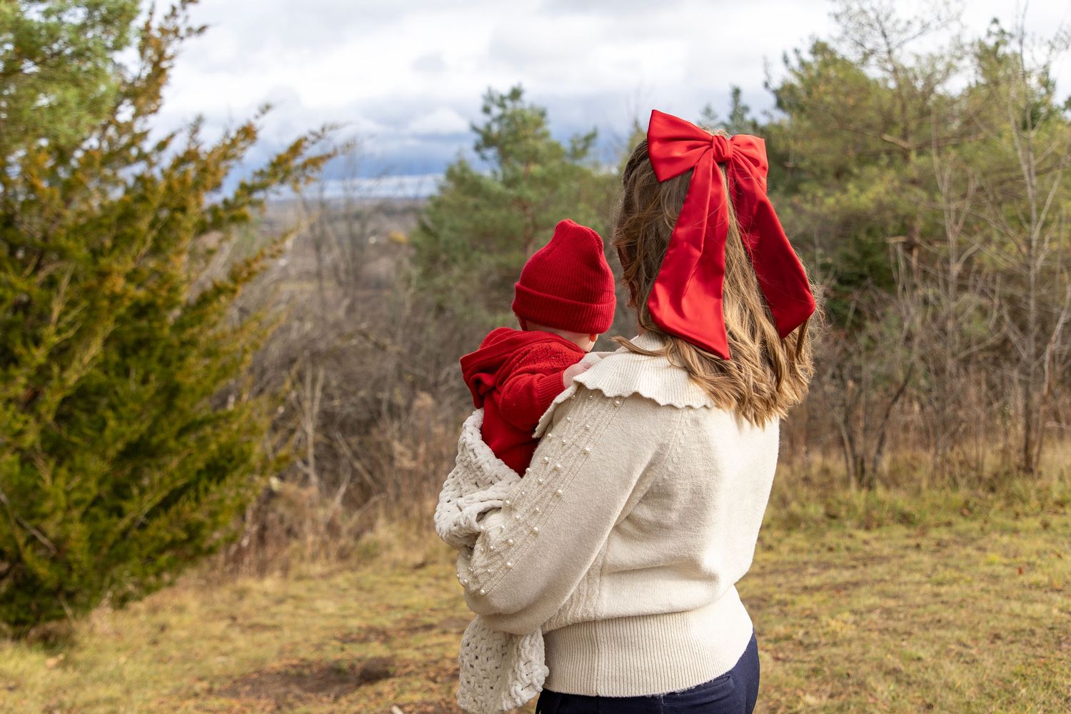 A mom holding her young baby boy both looking at the view in front of them. Mom is wearing a red bow and baby, a red hat