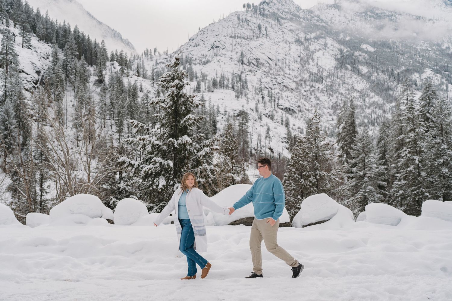 Couple walking through a snowy mountain landscape with evergreen trees and white peaks in the background. Leavenworth, WA