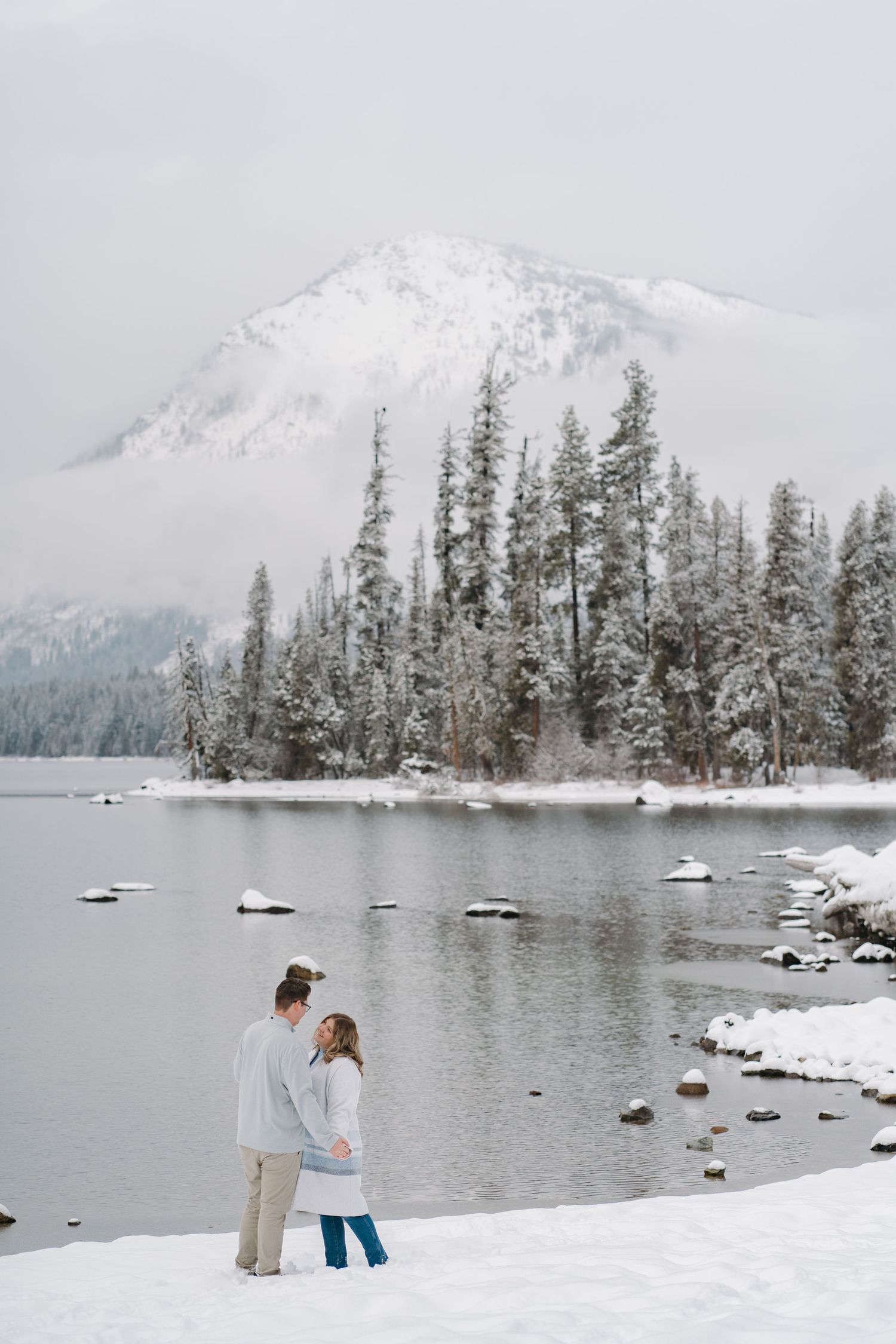 A couple stands together on a snowy shoreline with evergreen trees and mountains creating a serene winter landscape. Leavenworth, WA