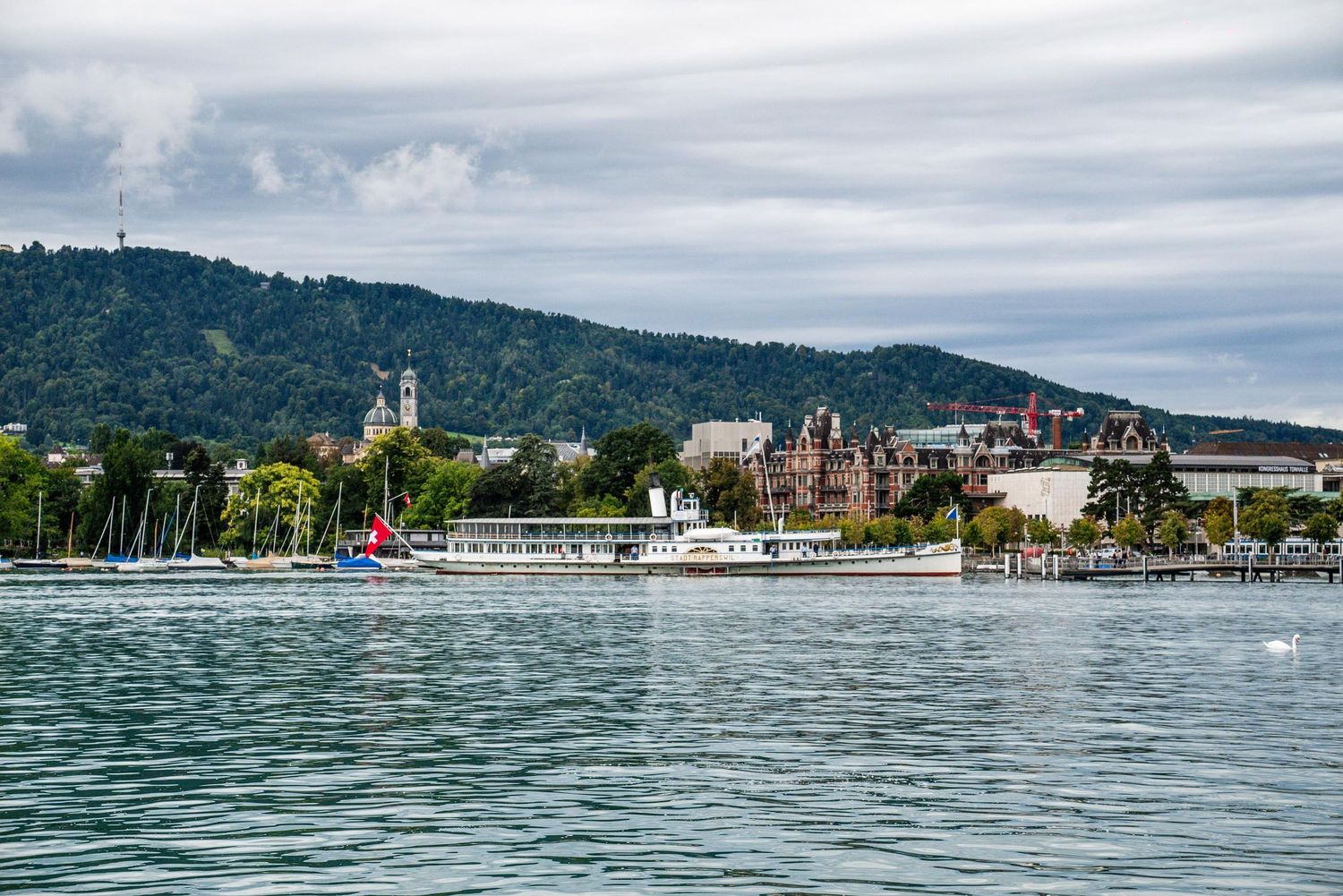 Panoramic view of Lake Worthersee in Austria with mountains, boats, and buildings along the shoreline under cloudy skies.