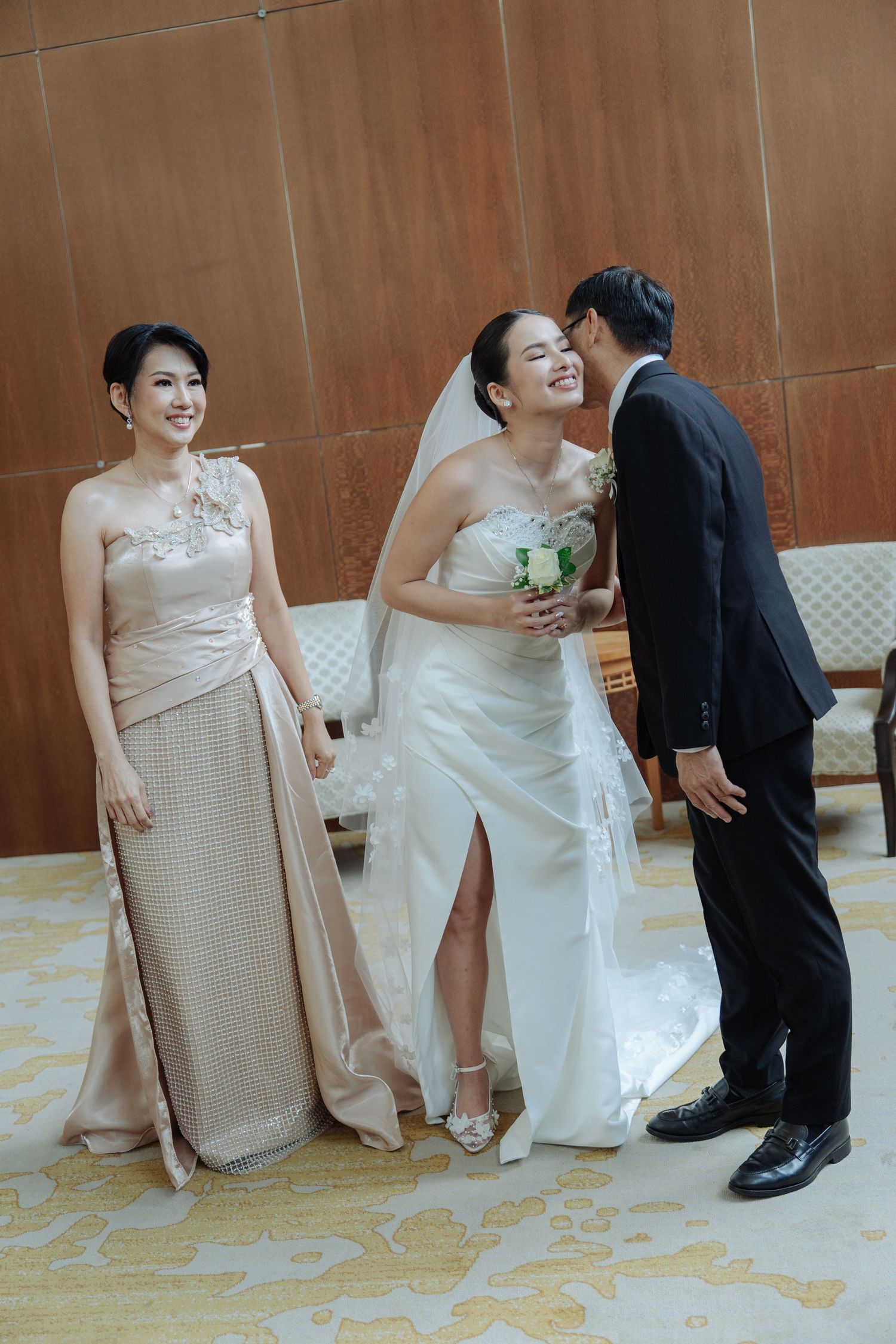 Groom whispering to smiling bride while mother of bride stands beside them in elegant venue.