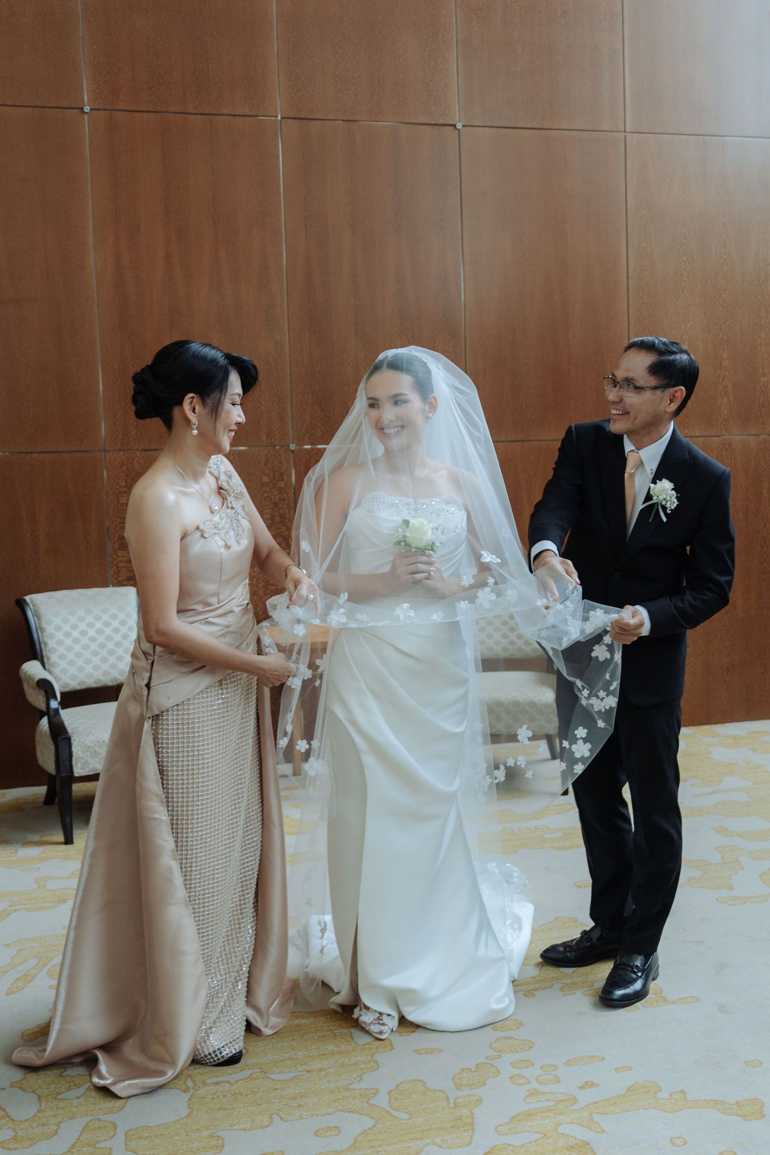 Bride smiling as parents help adjust her floral wedding veil in a warmly lit venue.