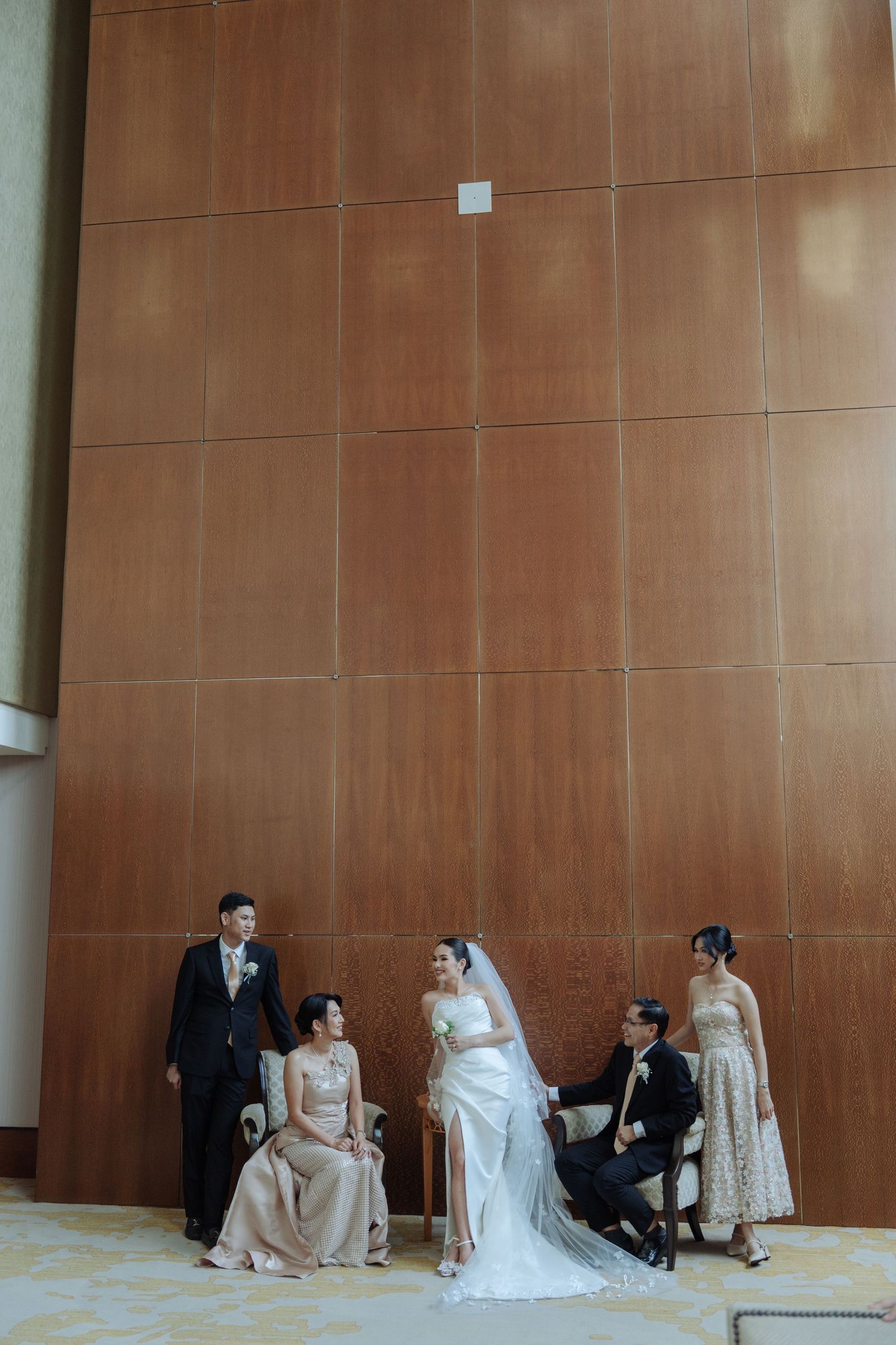 Wedding party poses elegantly against a wooden wall, with bride and groom centered among family members in formal attire.