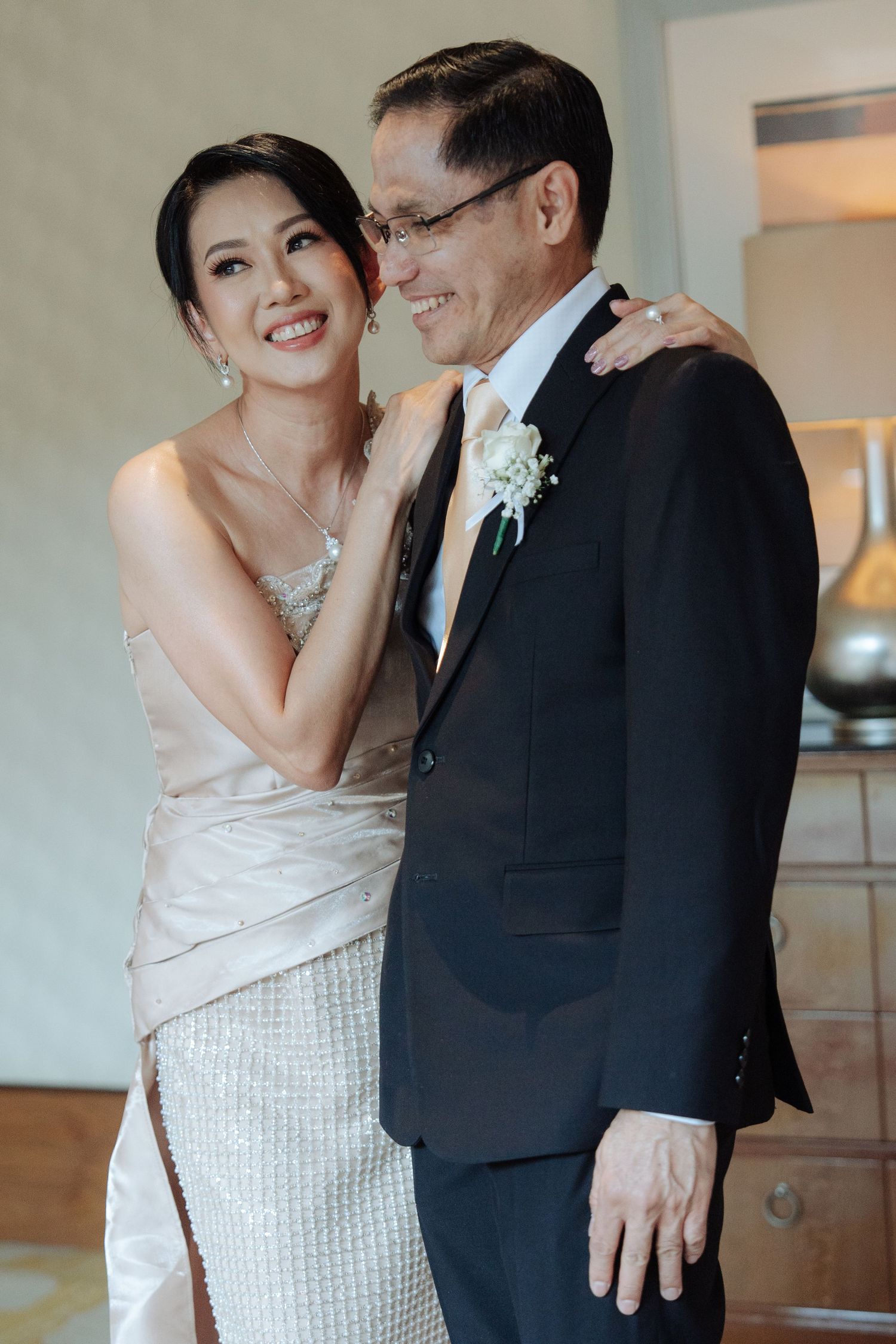 Smiling bride in champagne dress embraces older man in black suit with boutonniere at wedding celebration.