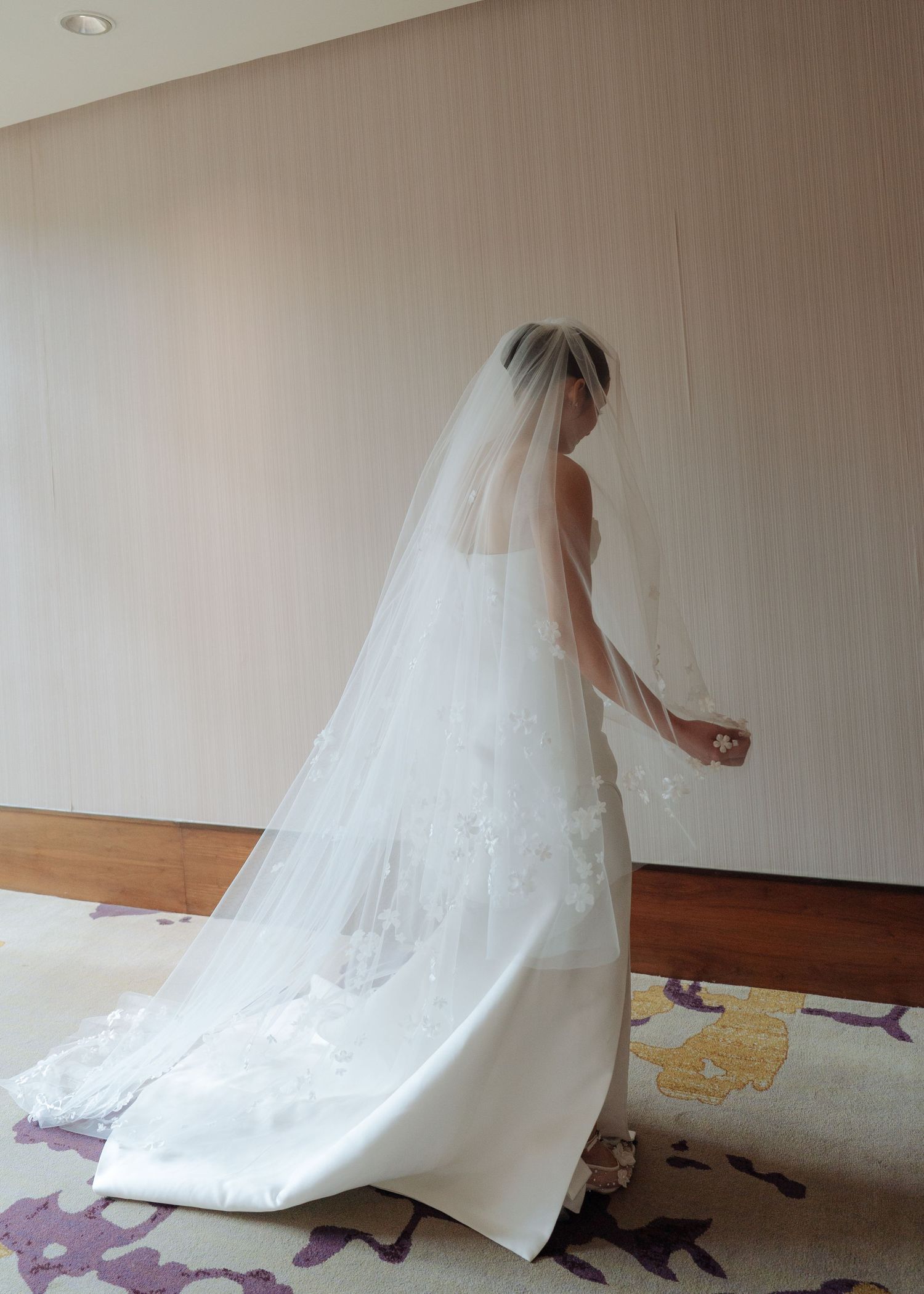 Bride in white strapless gown with long floral cathedral veil stands gracefully in hotel wedding suite.