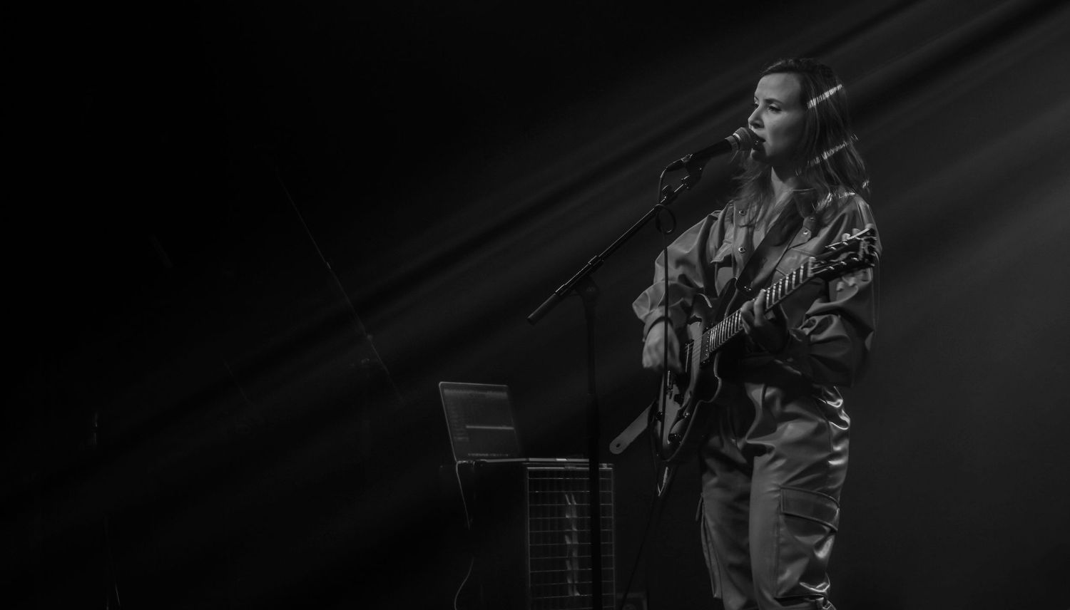 A black and white photograph of a guitarist performing on stage with dramatic lighting and shadows.