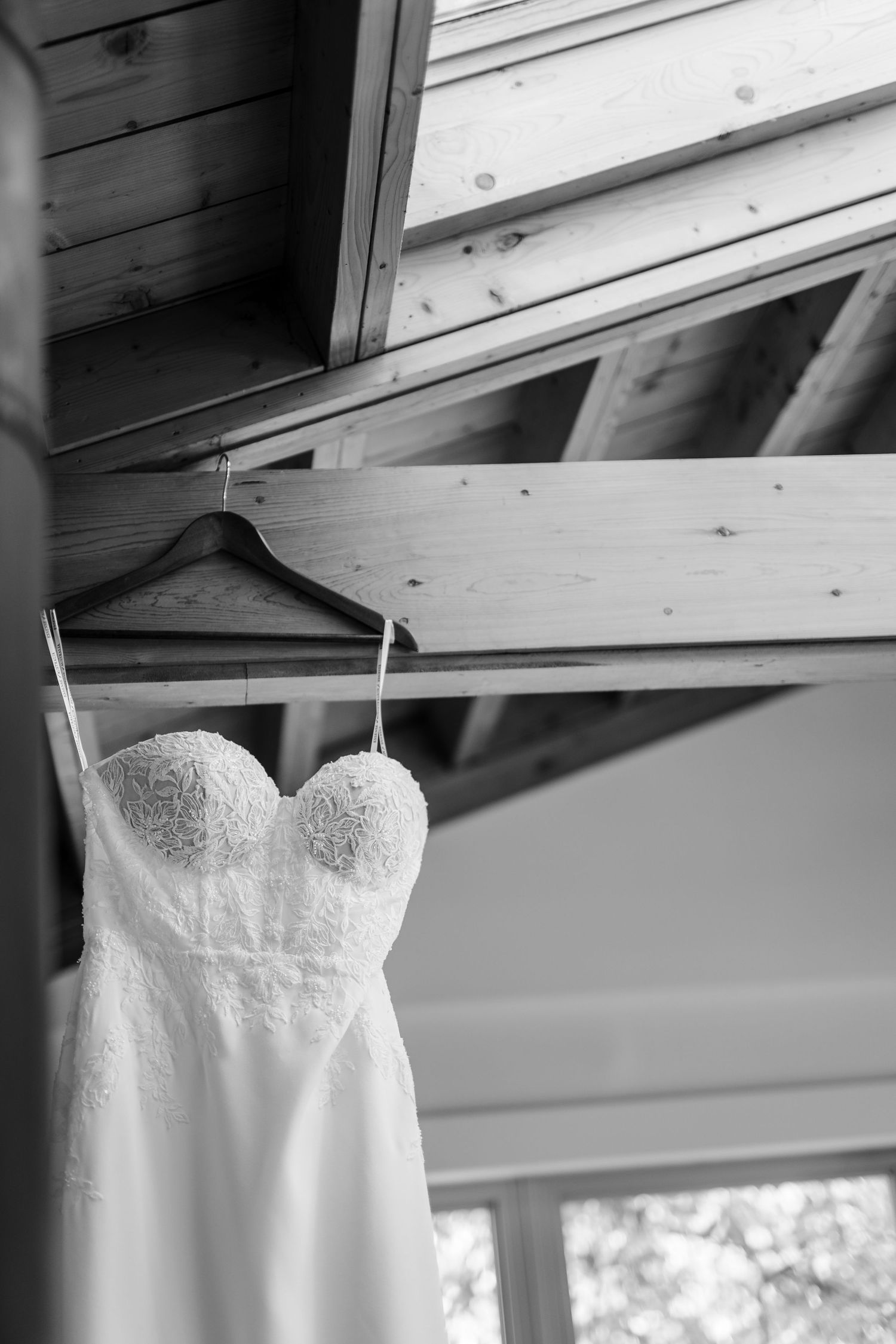 A white lace wedding dress hangs from a wooden beam in natural lighting, showcasing its strapless sweetheart neckline.
