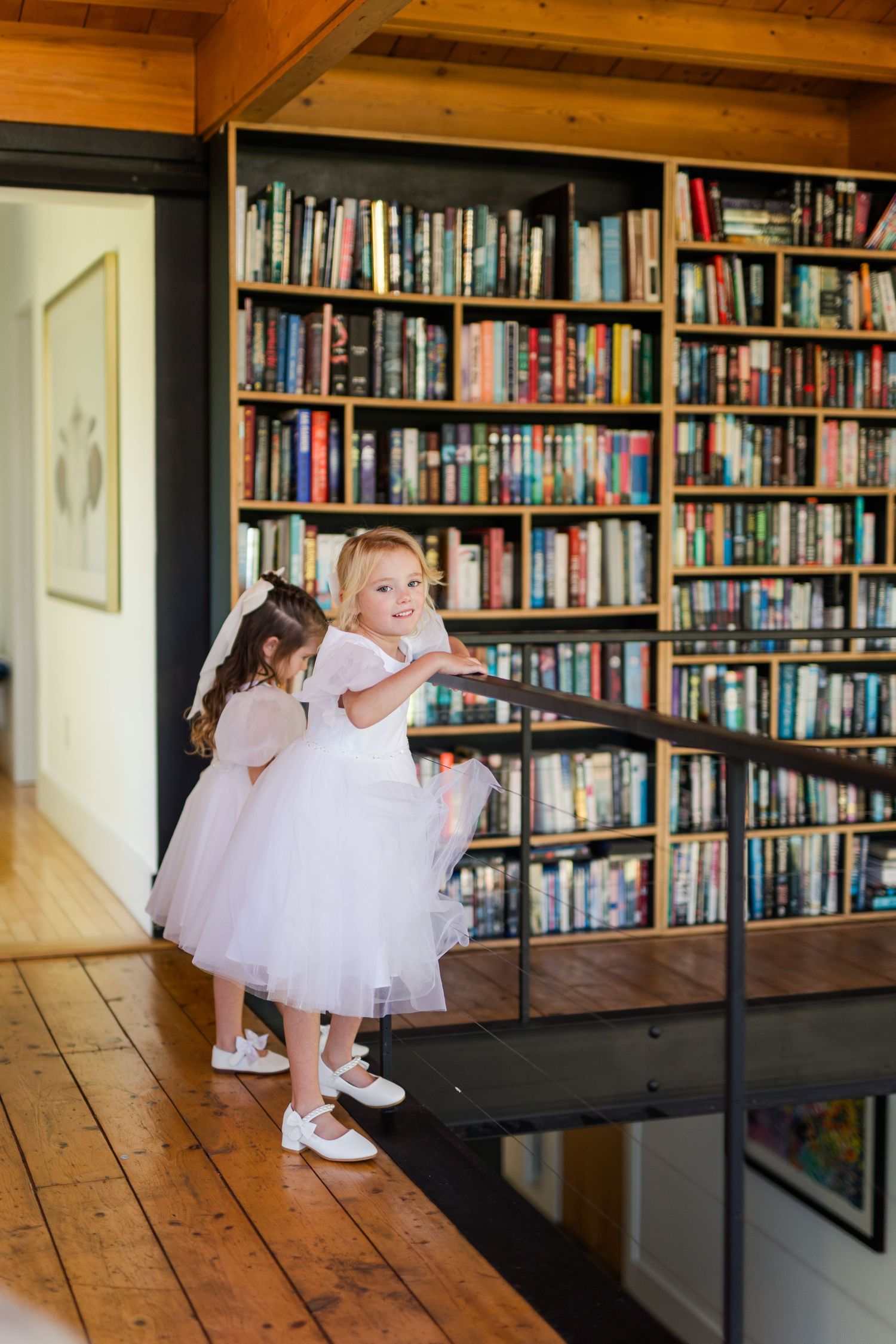 Two children in white tulle dresses explore a library with floor-to-ceiling bookshelves and wooden floors.