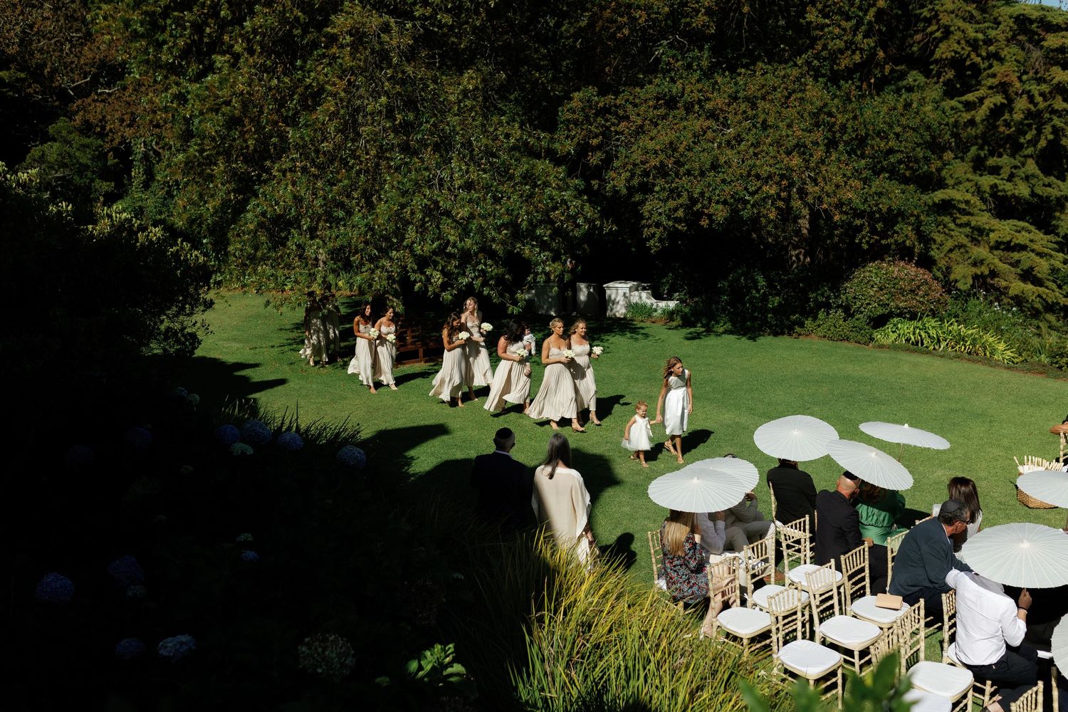 Wedding ceremony guests seated in white chairs on a lush green lawn surrounded by trees and gardens.