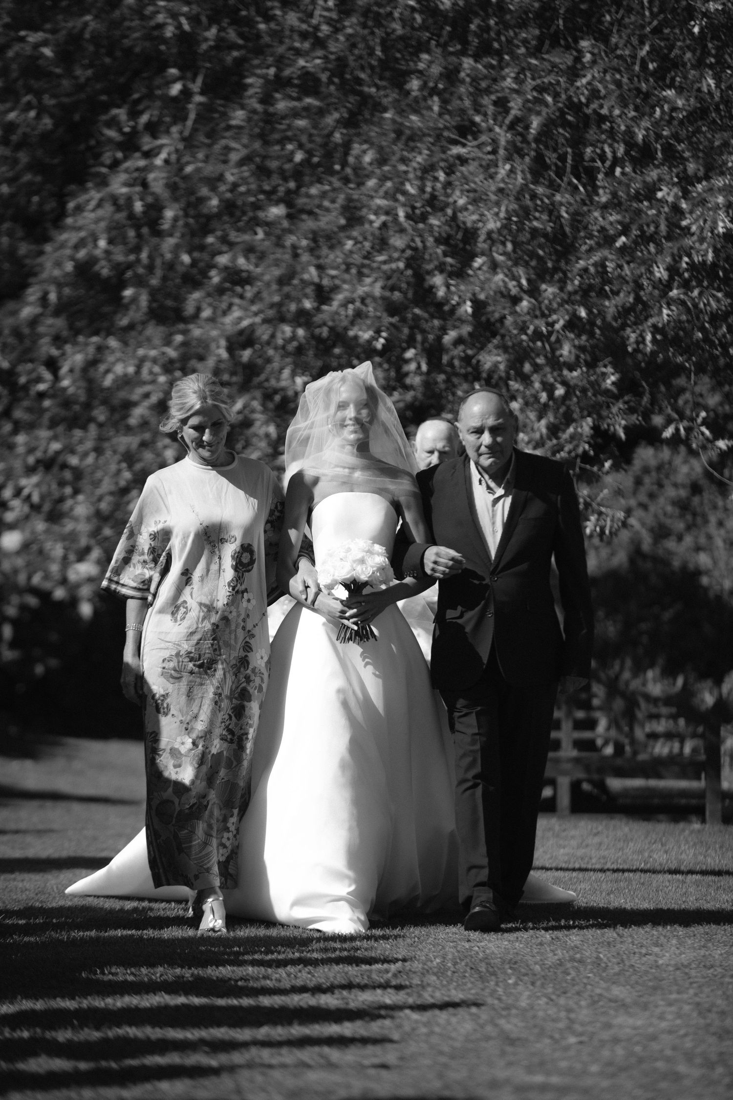 Black and white photo of a traditional wedding procession walking outdoors along a garden path.