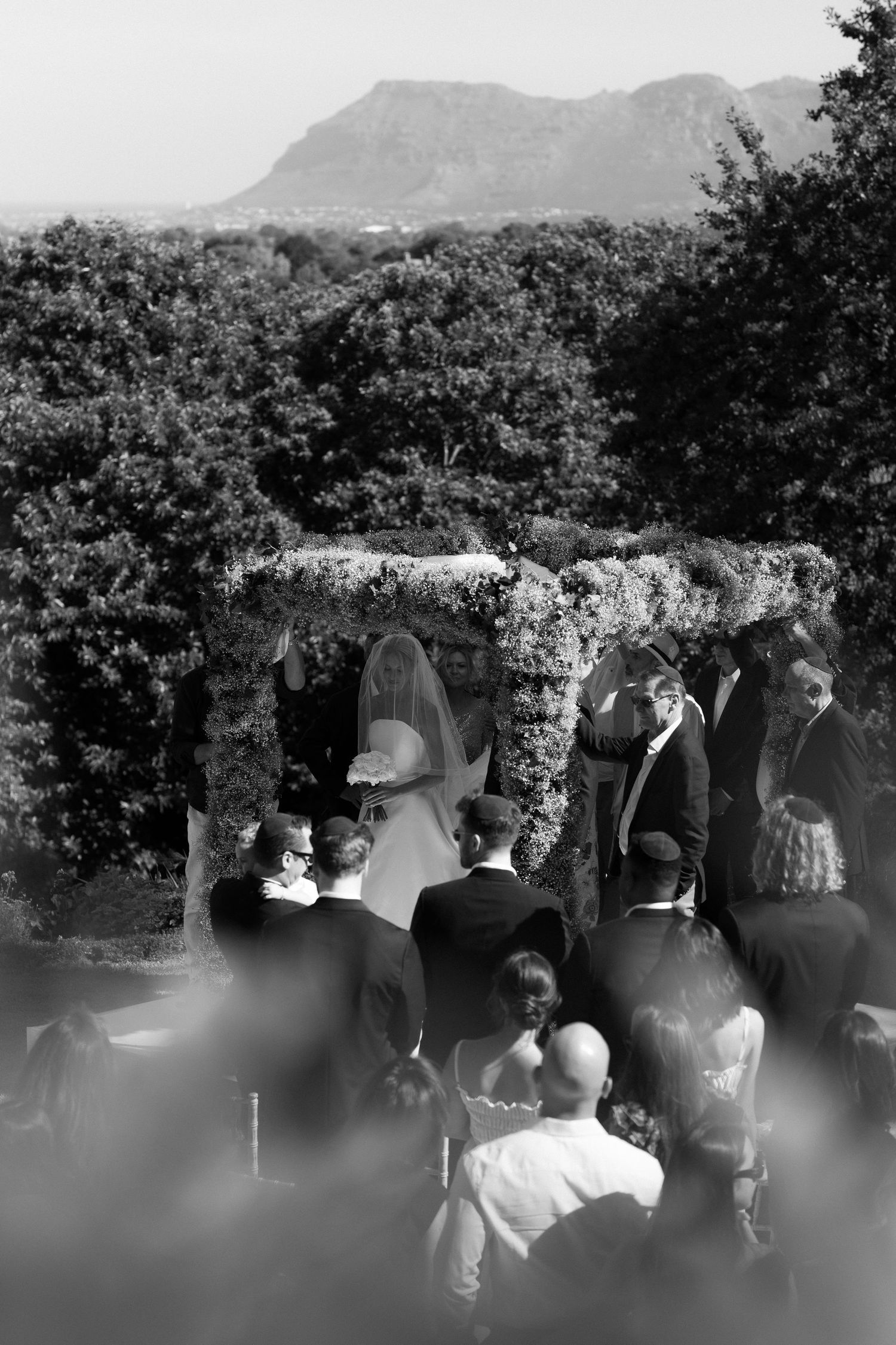 Wedding ceremony guests gather outdoors with mountains and lush greenery in the background.