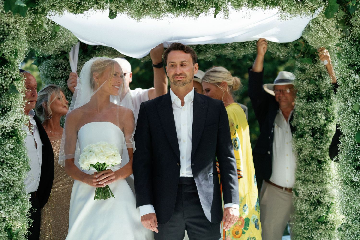 A wedding party poses outdoors under lush green foliage during a summer celebration.