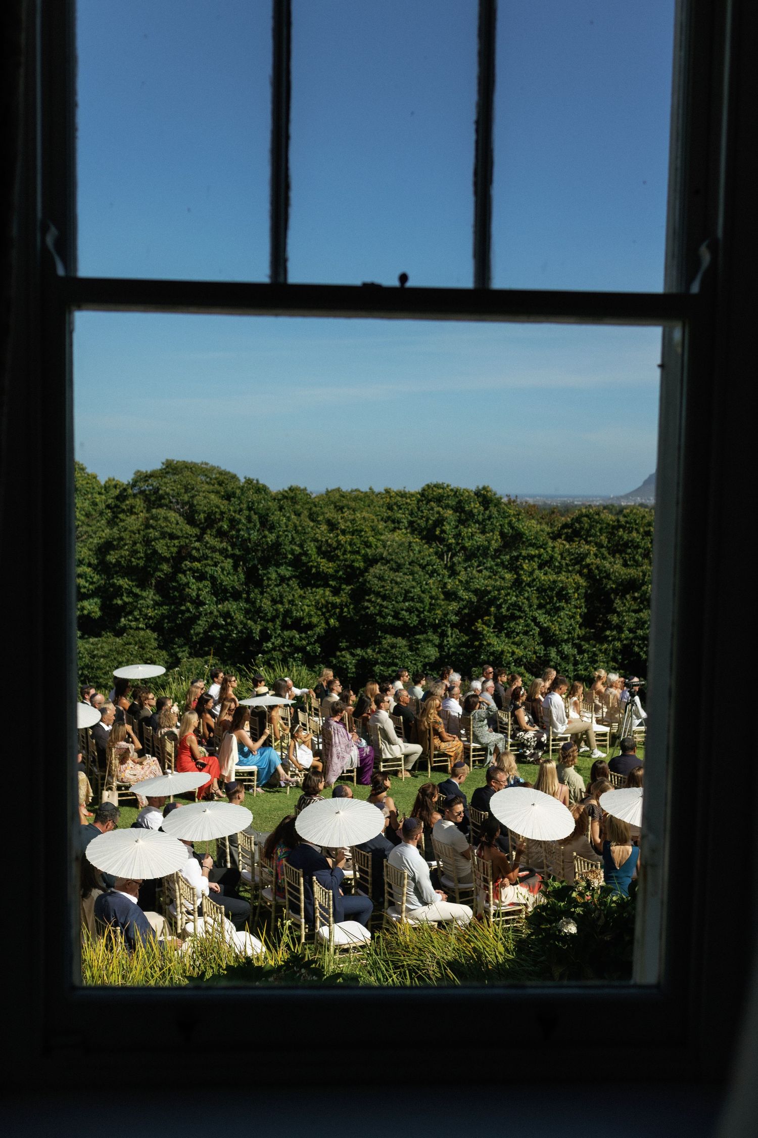 Wedding guests with white parasols gather outdoors for a sunny ceremony viewed through a window frame.