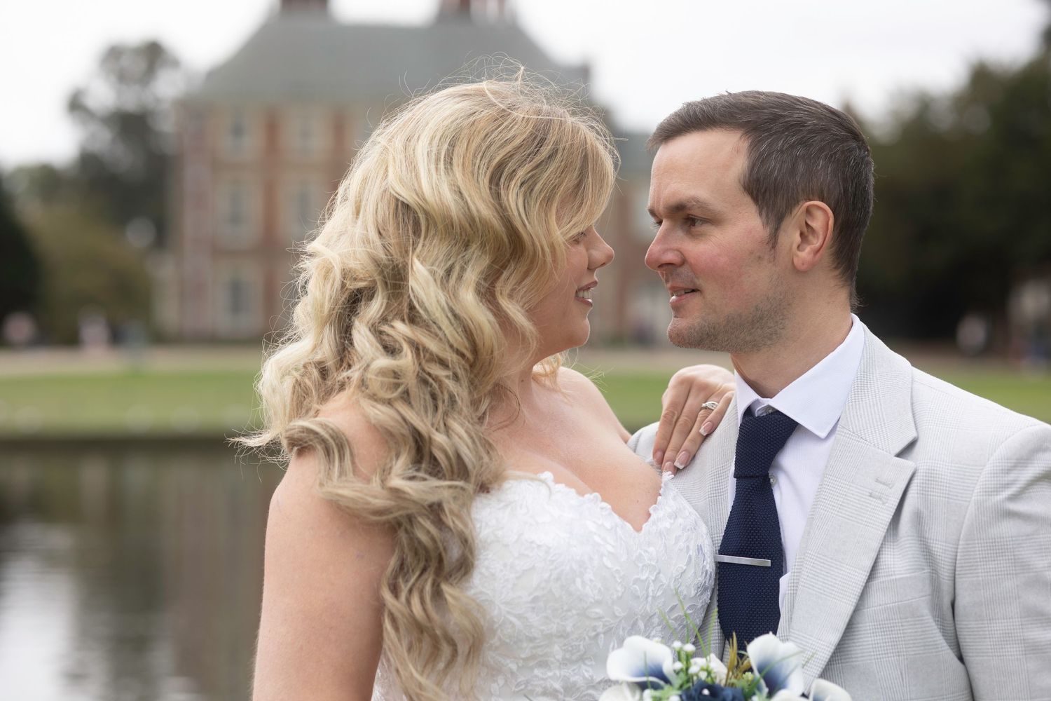 A romantic wedding couple shares an intimate moment by a lake with a stately brick building in the background.