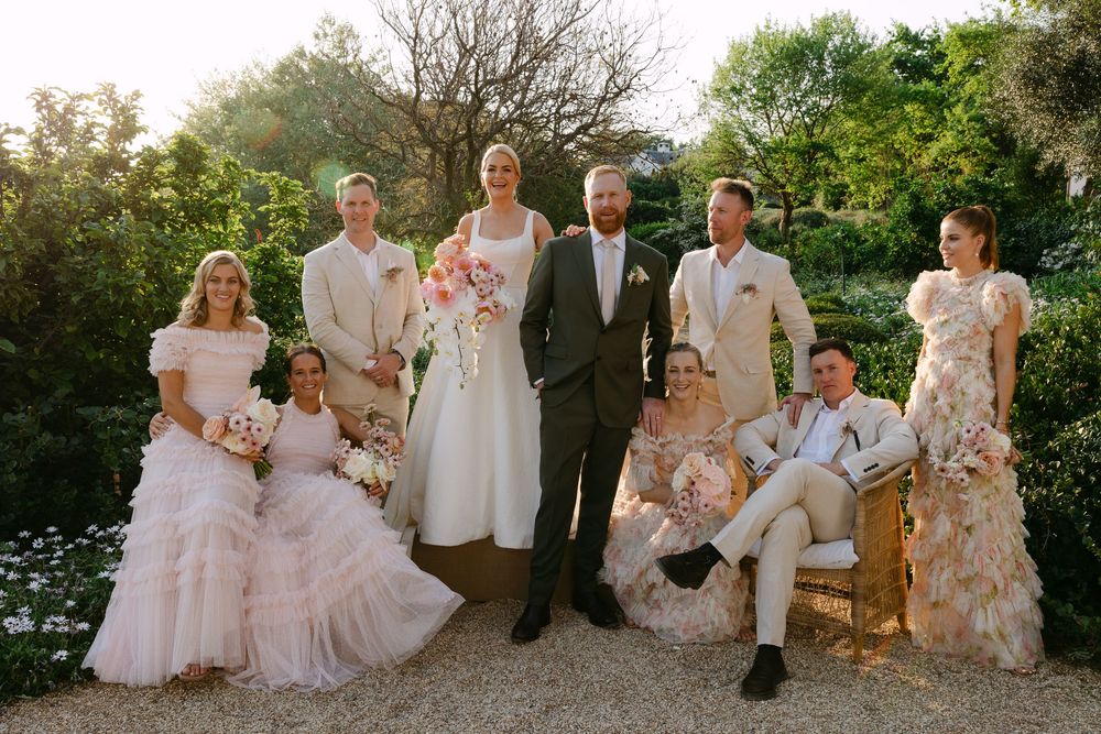 A wedding party in soft pastel attire poses together in a garden setting during golden hour lighting.