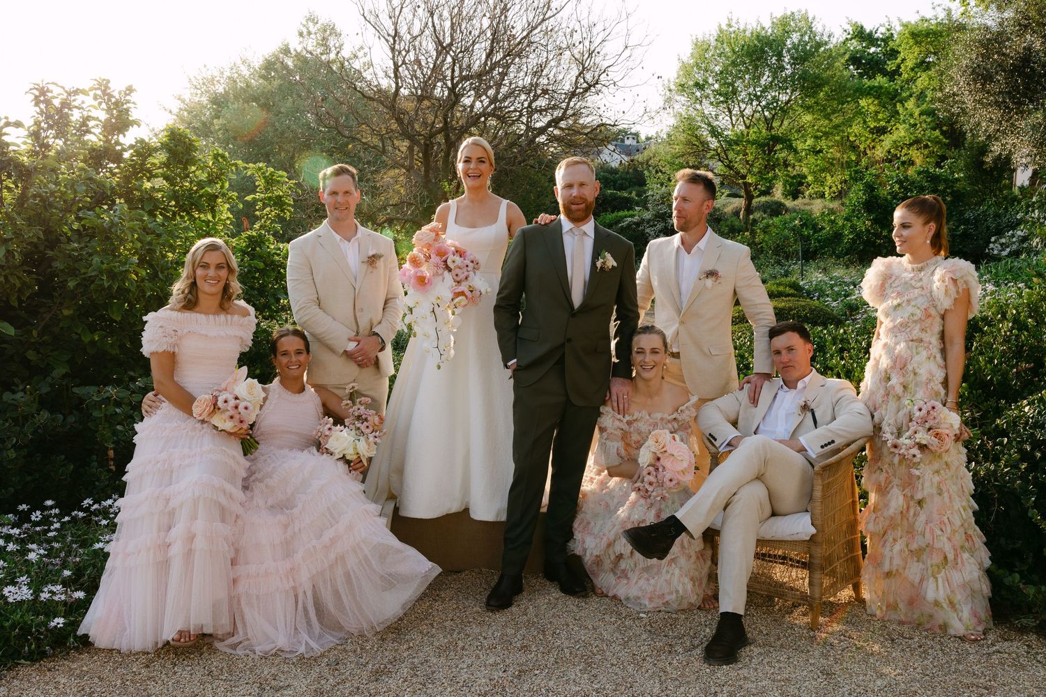 A wedding party in soft pastel attire poses together in a garden setting during golden hour lighting.