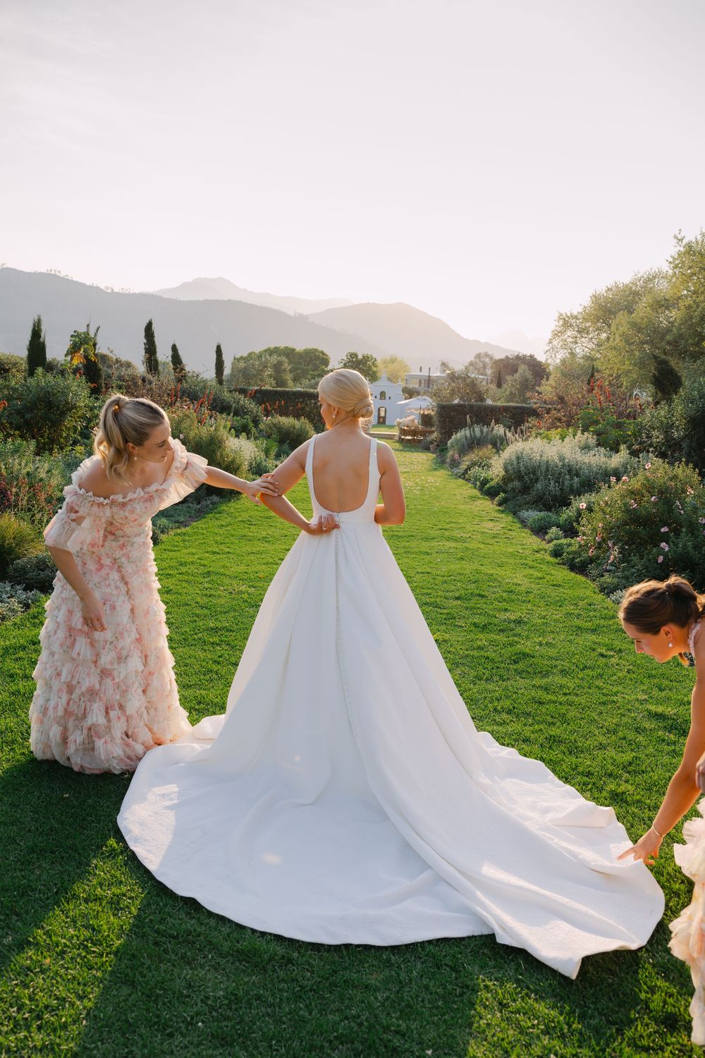 A wedding dress train being arranged on lush green grass with mountains and cypress trees in the background at sunset.