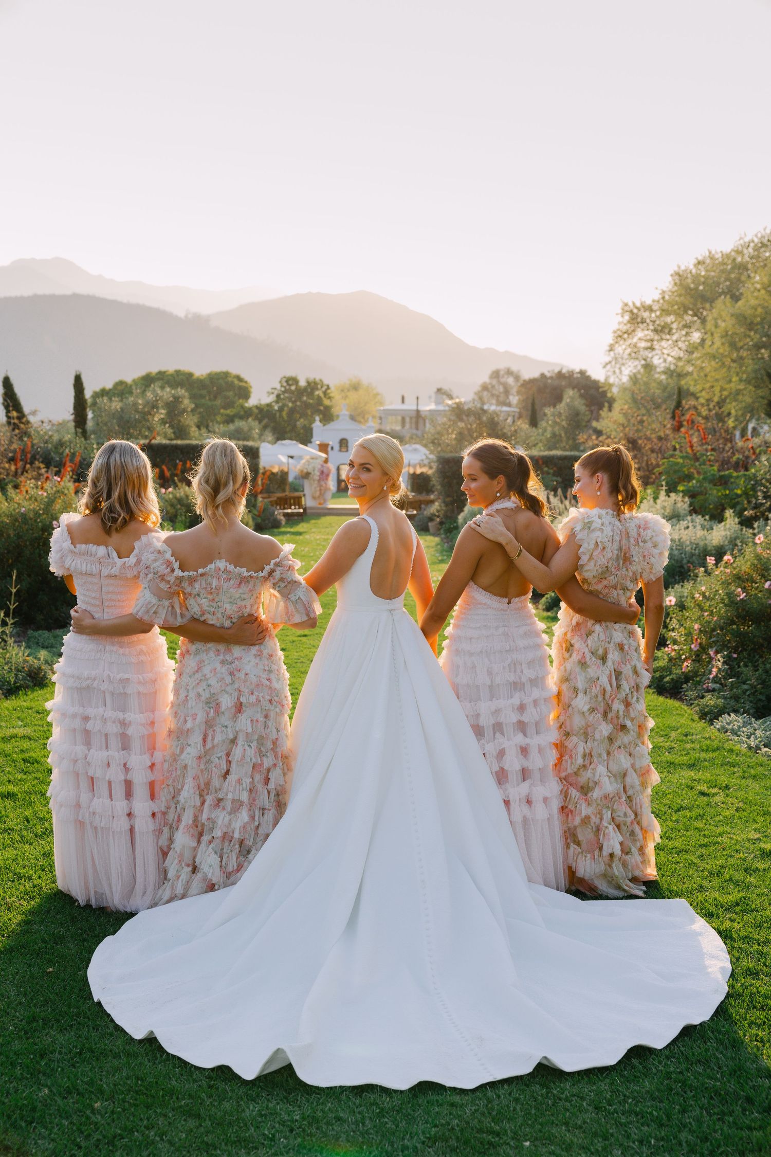 A bride and bridesmaids in pink floral dresses pose together on a grassy hill at sunset.