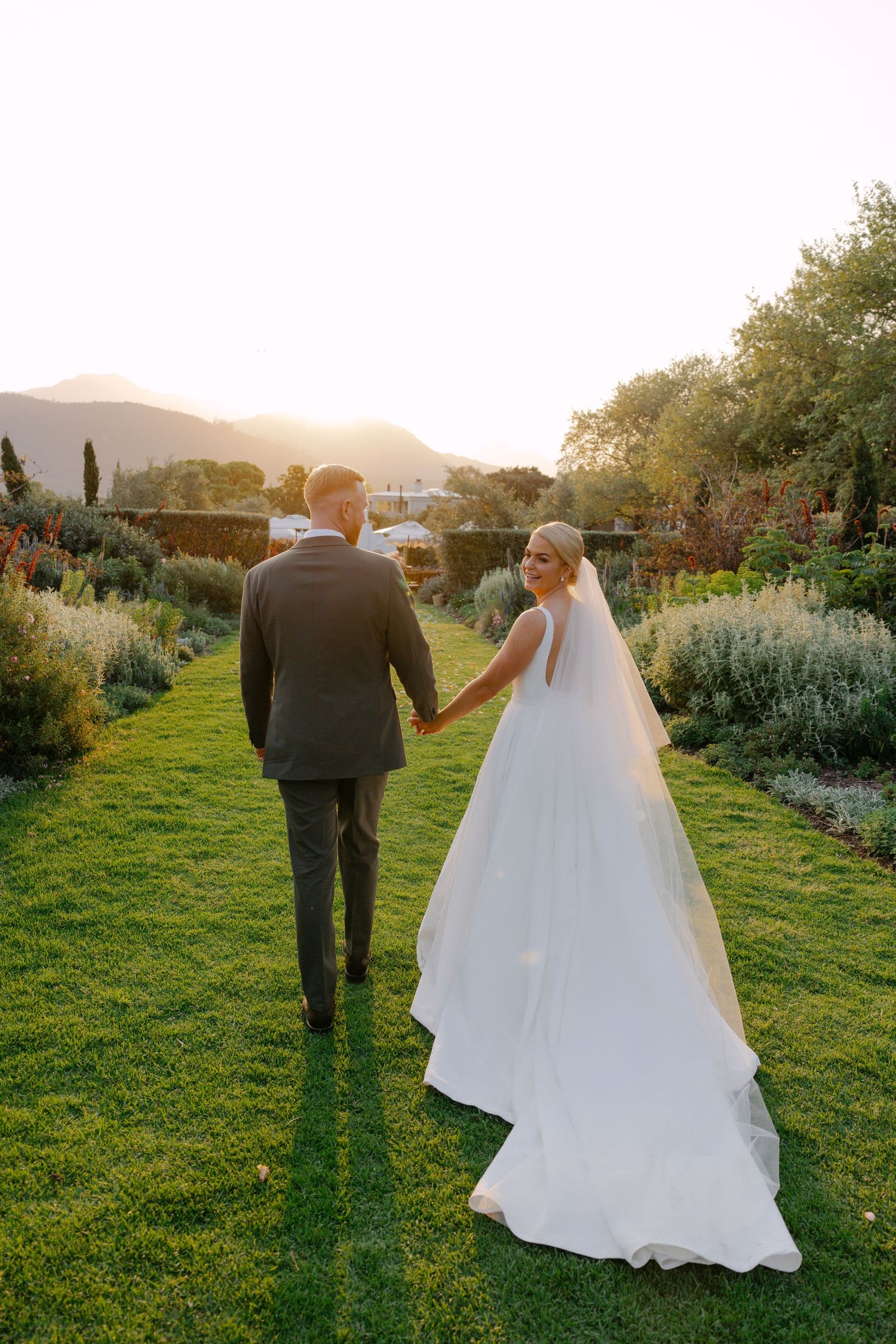 A couple walking through a garden path with mountains in the distance as the bride's white dress trails behind them.