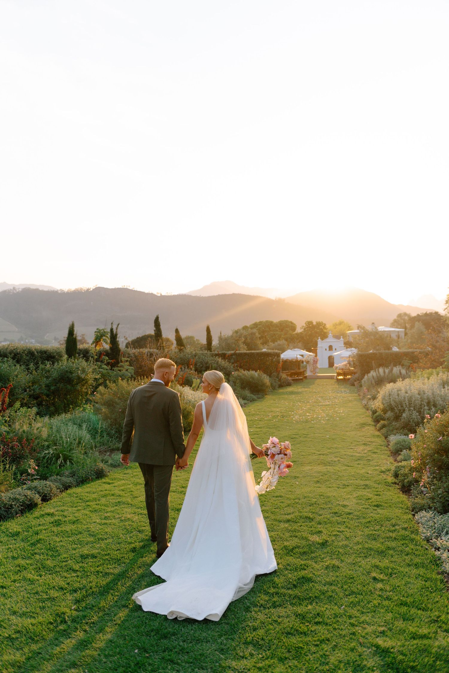 Couple walking through a lush garden path at sunset with long shadows cast across the green grass.