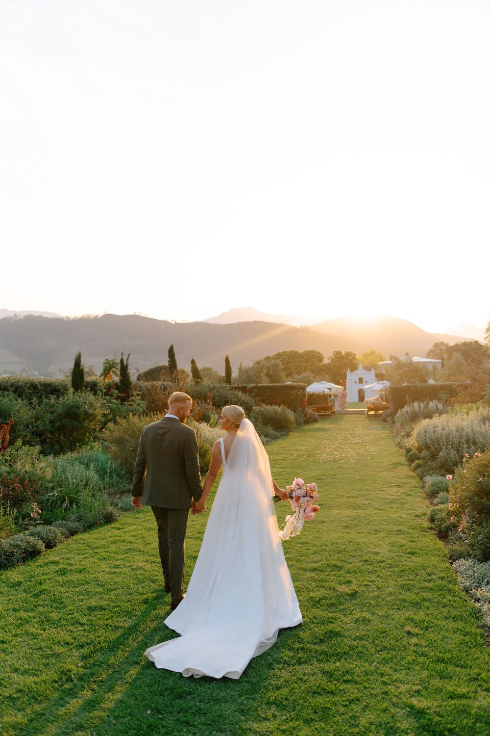 Couple walking through a lush garden path at sunset with long shadows cast across the green grass.