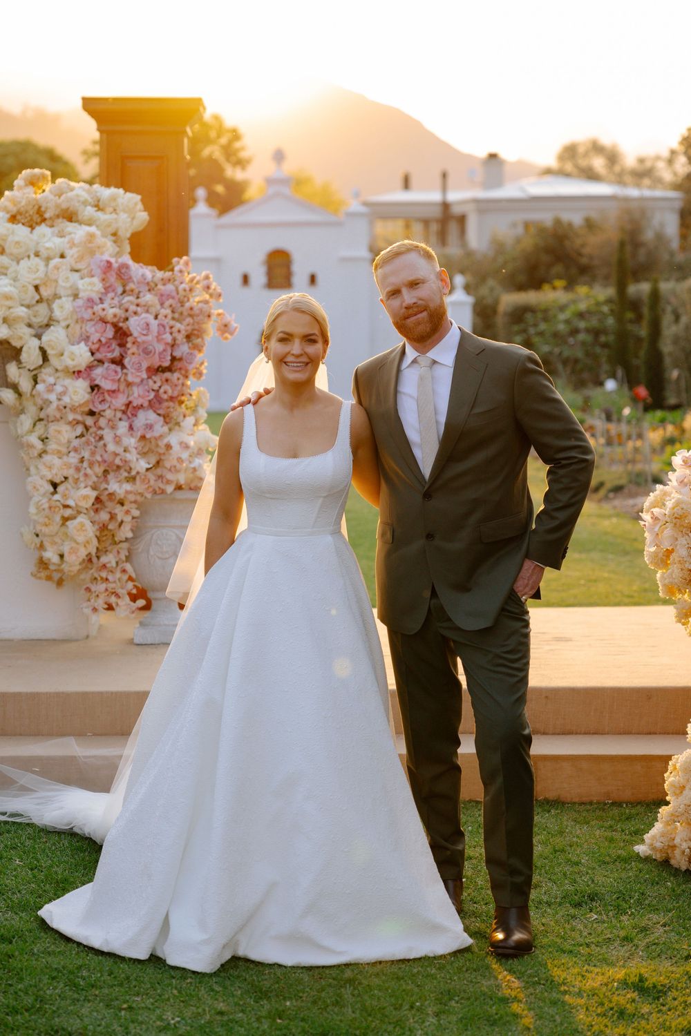 A wedding couple poses in a garden setting during golden hour with pink and white floral arrangements.
