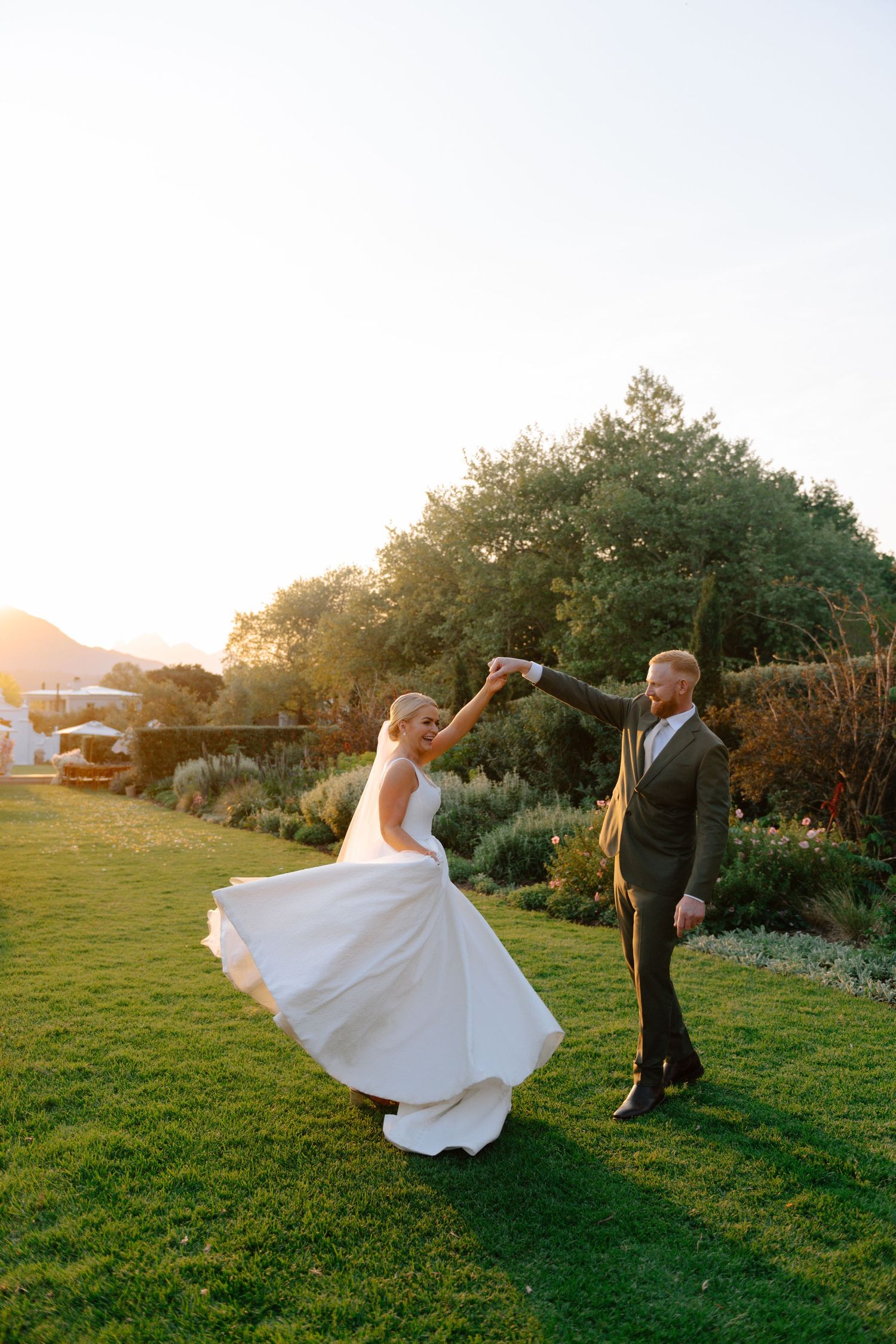 A joyful wedding couple dances together on a lawn at sunset surrounded by lush gardens.