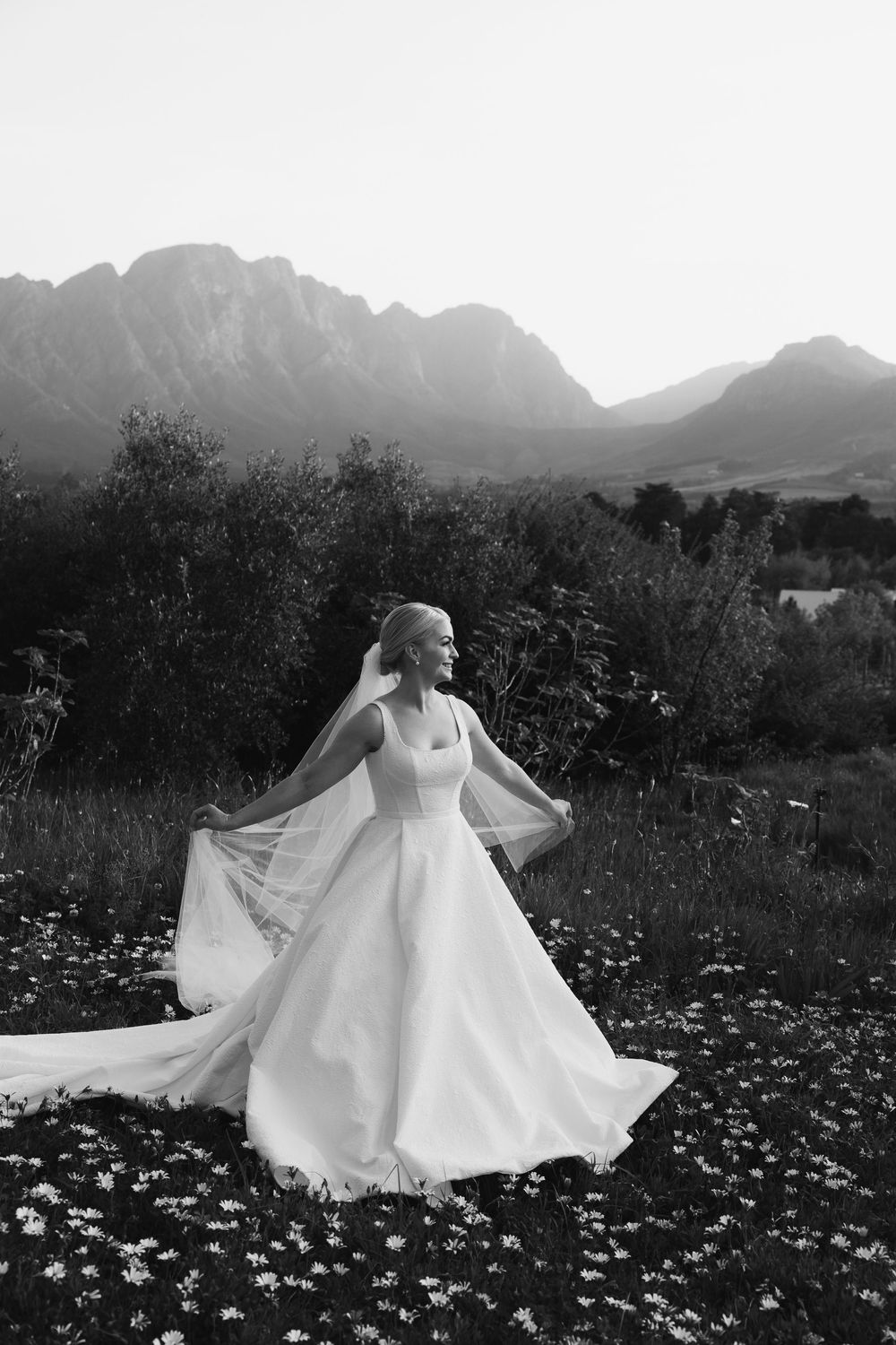 A bride in a flowing white dress twirls among wildflowers with majestic mountains in the background.