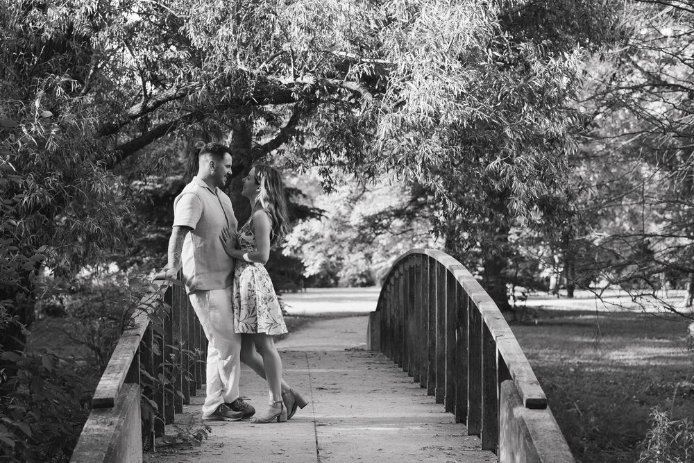A couple stands together on a curved footbridge in a scenic park setting captured in black and white.