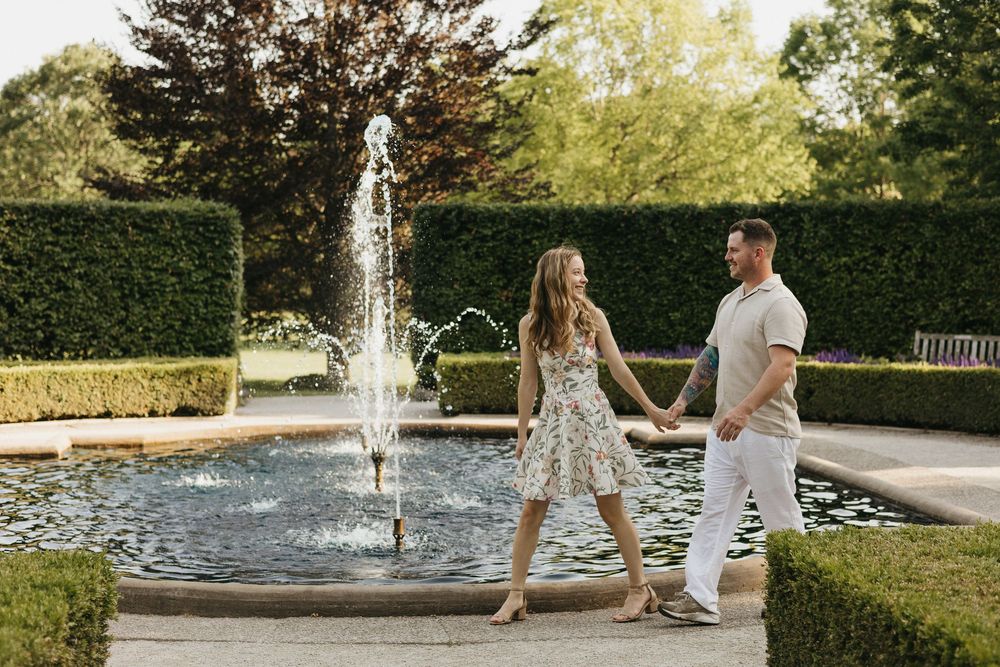A couple sits together near a garden fountain on a sunny summer day while holding hands.