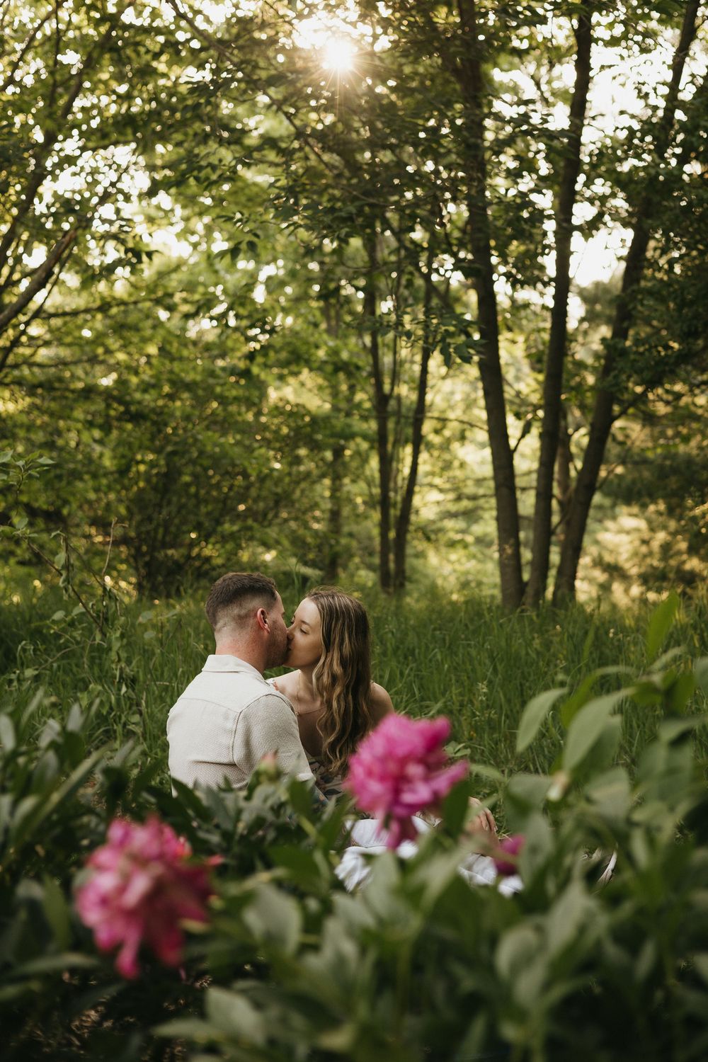 A romantic couple shares a tender moment in a sunlit forest with pink flowers in the foreground.
