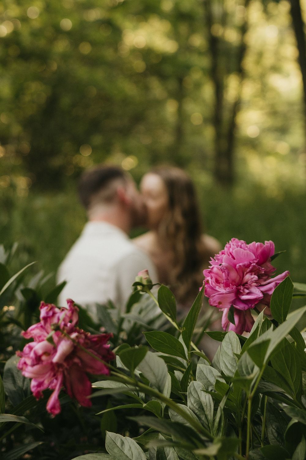 Pink peonies bloom in foreground while couple shares intimate moment in blurred forest background.