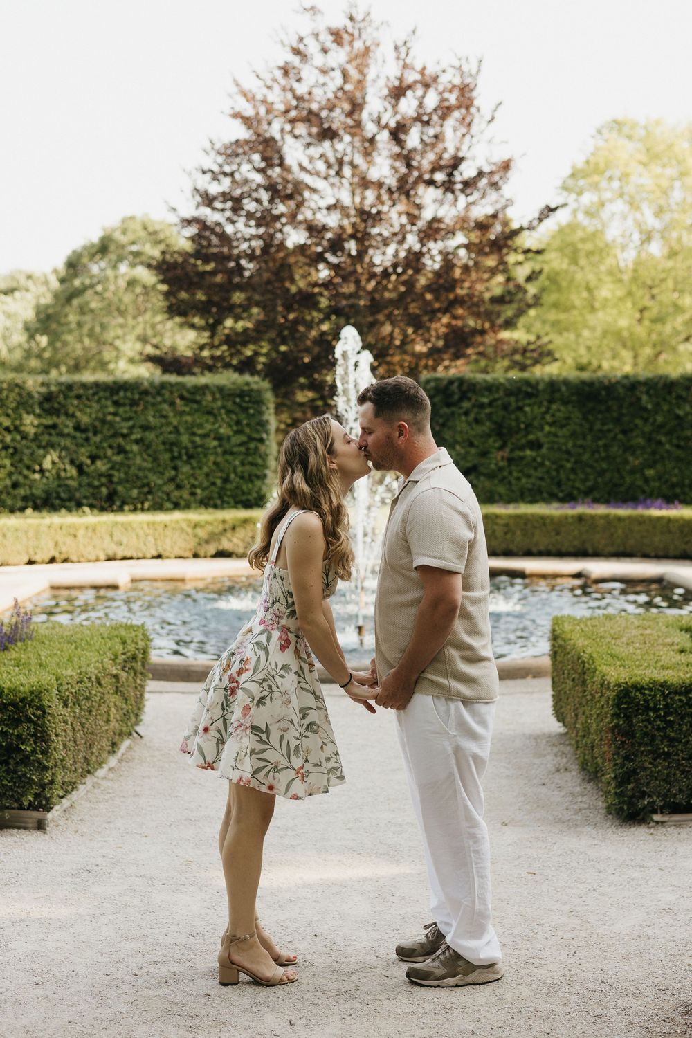 A couple shares a romantic moment in a garden pathway surrounded by manicured hedges on a sunny day.