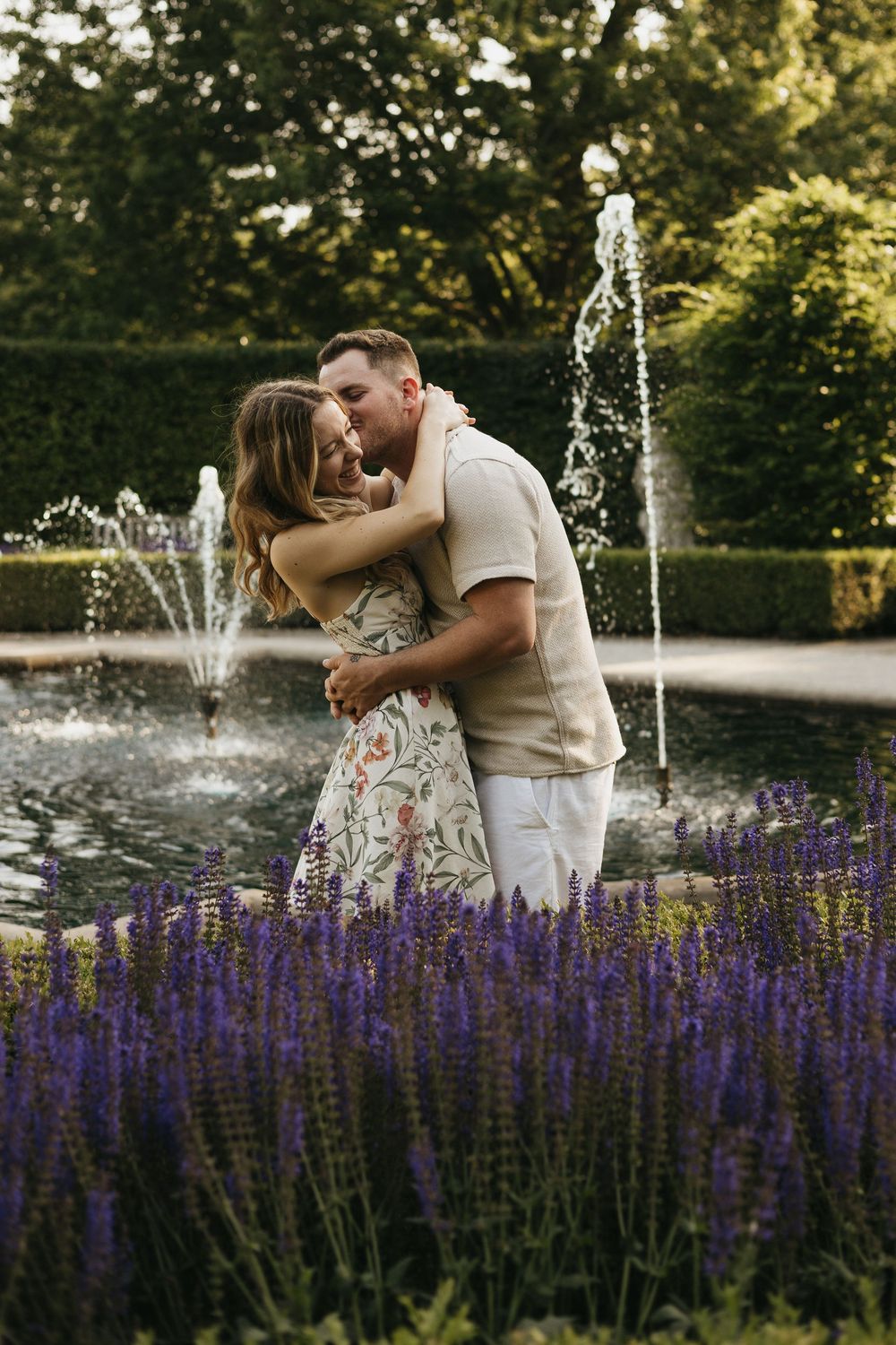 A couple embraces near a fountain with vibrant purple lavender flowers blooming in the garden.