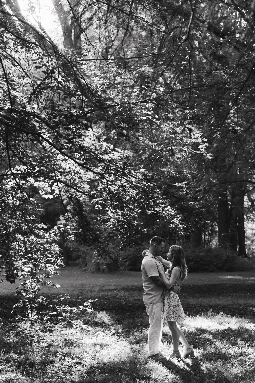 Black and white photo of two people walking together in a sunlit wooded area with dappled light filtering through trees.