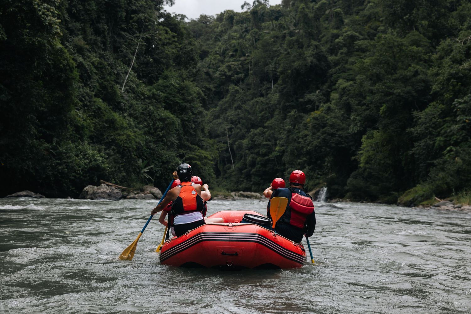 Three people paddling a red inflatable raft through calm river waters surrounded by forest.
